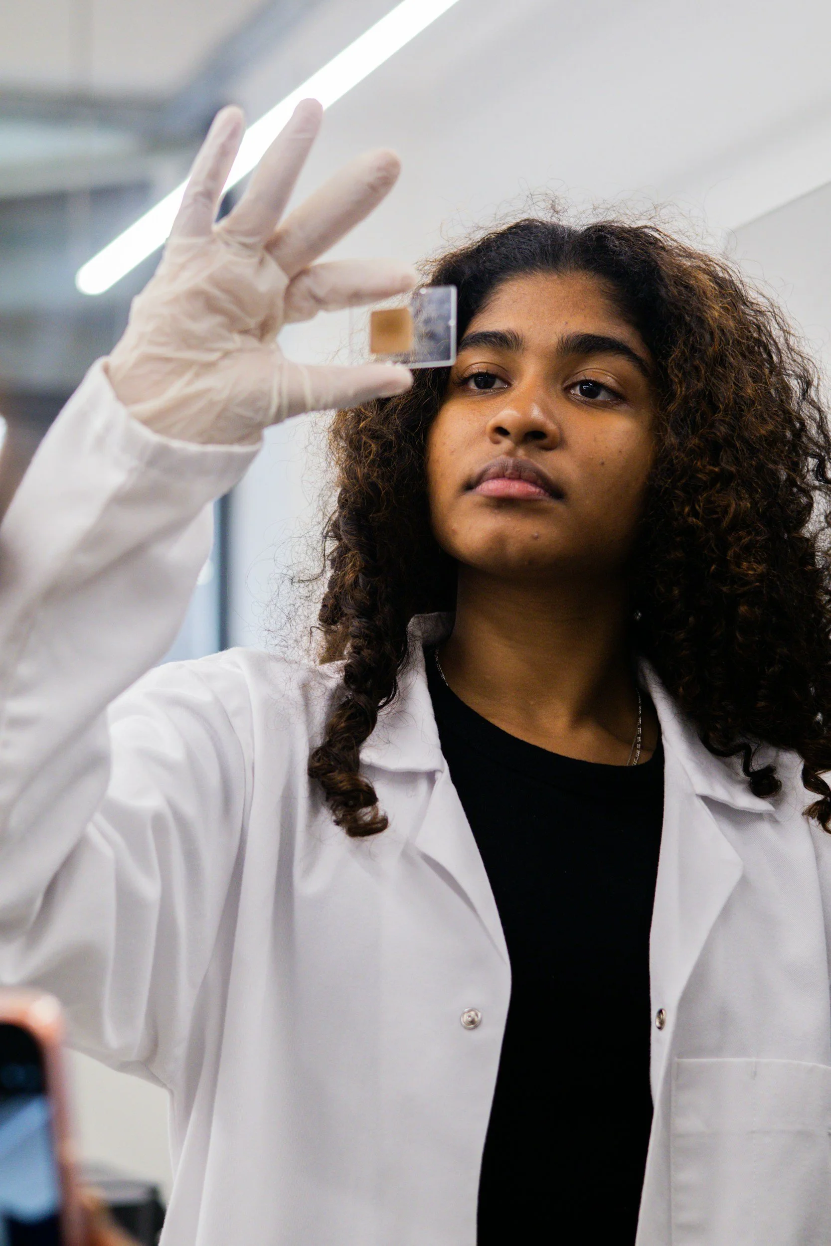 A woman with curly hair wearing a white lab coat and white gloves holds a small square object close to her eye level. The background shows a well-lit indoor space with fluorescent lighting.