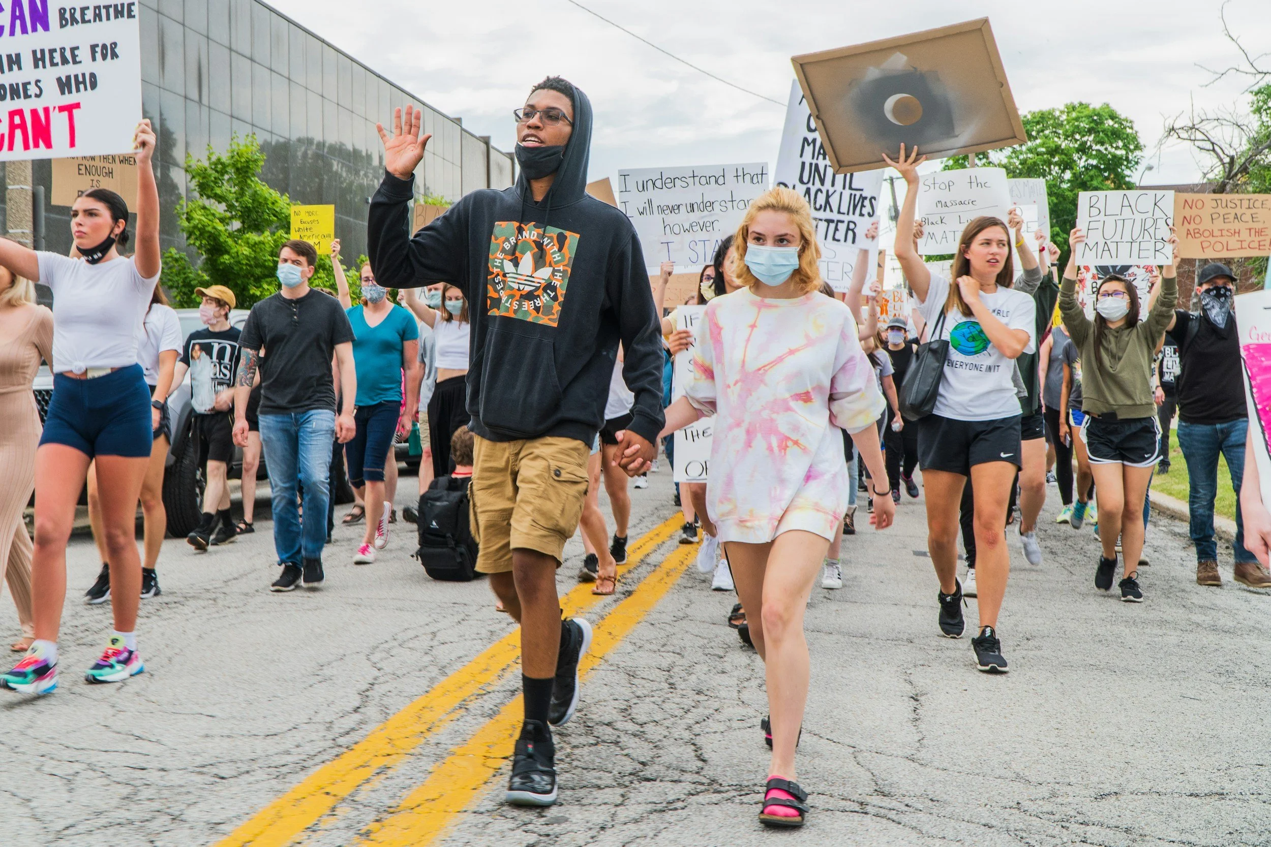 A diverse group of people participating in a protest march during daytime, holding signs advocating for racial justice, with some wearing masks. The crowd is walking on a cracked street, surrounded by trees and buildings, with cloudy sky overhead.