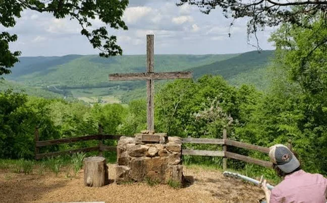 Wooden cross mounted on a stone pedestal overlooking green hills, with a person wearing a pink shirt and gray cap reading a book nearby.