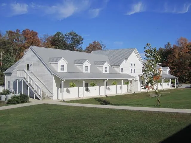 A white barn-style building with multiple dormer windows, a metal roof, and a wrap-around porch, set against a backdrop of trees with fall foliage under a blue sky.