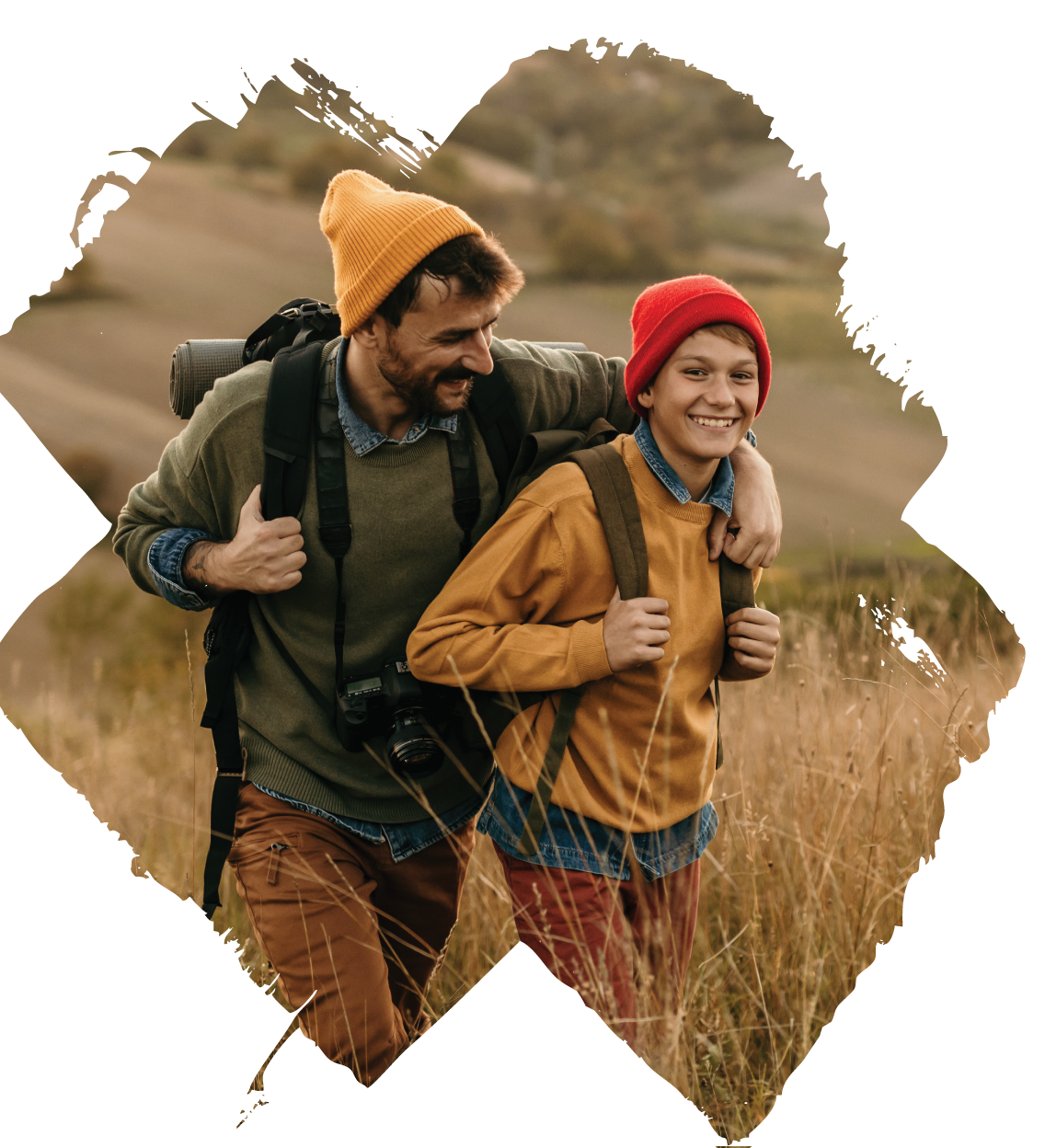 A man and a boy hiking outdoors through a grassy field, both wearing red and yellow beanies and backpacks, smiling and enjoying the walk.