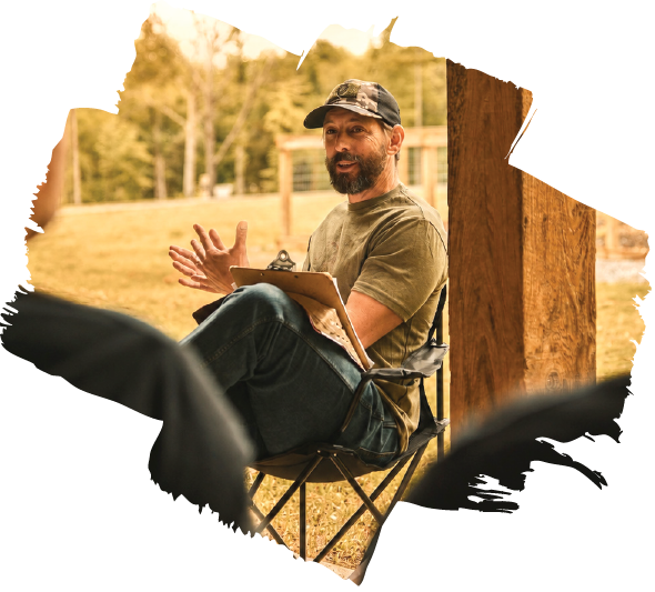 A man with a beard and cap sitting outdoors on a folding chair, holding a clipboard and speaking to people in an outdoor setting with trees and a grassy area.