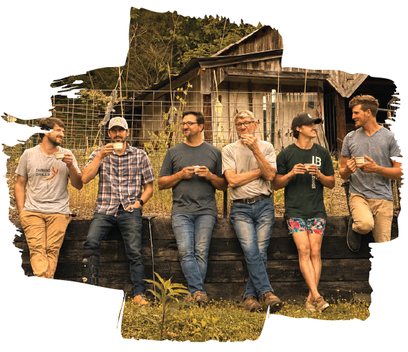 Group of six people outdoors, leaning against a wooden fence near a rustic barn, engaged in conversation and looking at their phones or drinking beverages.
