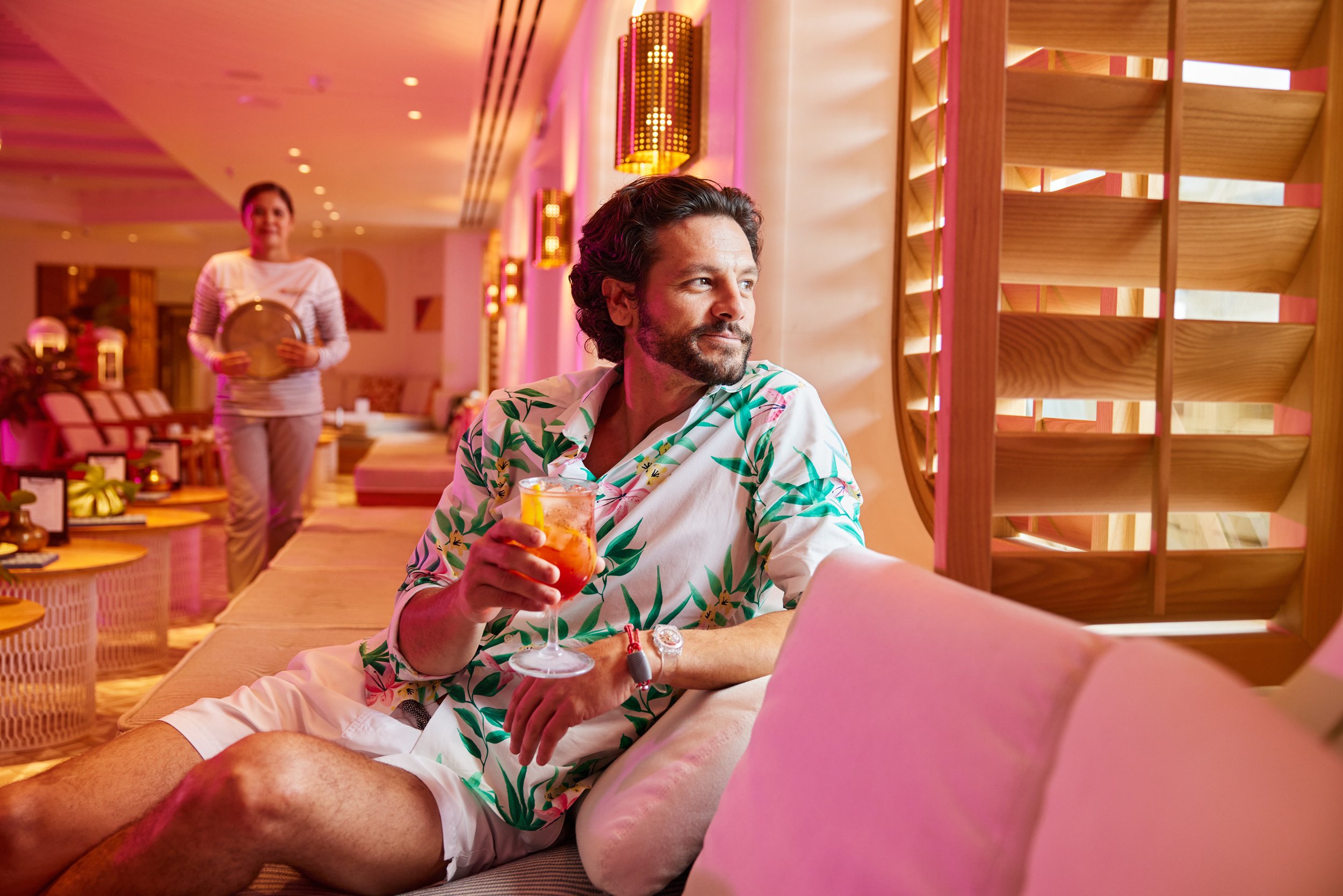 A man sitting at a table in a brightly lit restaurant, holding a colorful cocktail, with a woman walking towards him in the background holding a tray. The restaurant has modern decor with pink and purple lighting.