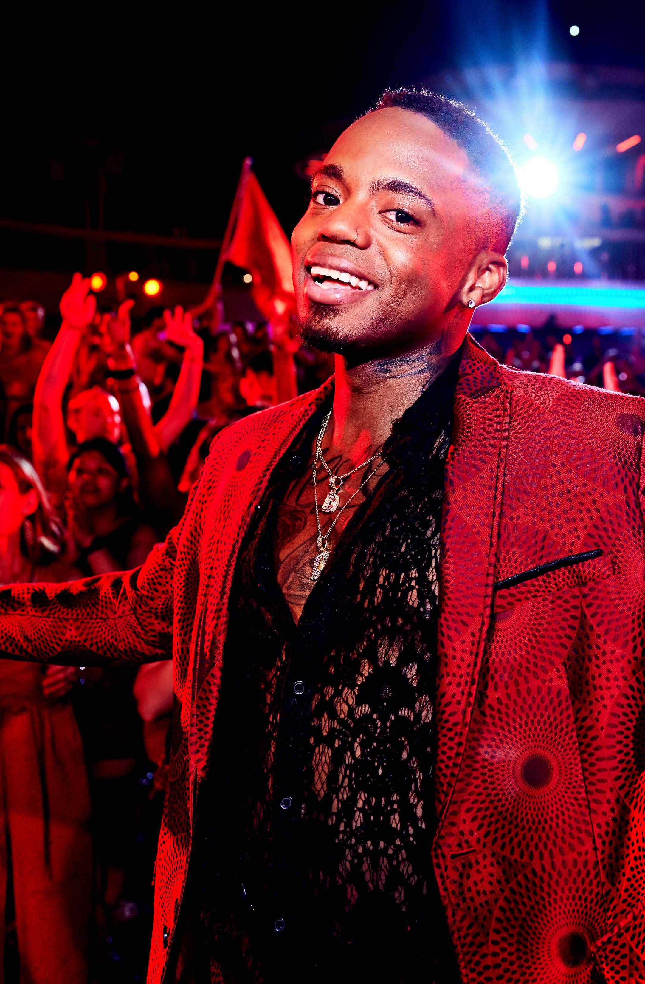 A smiling person with a short haircut, earrings, and layered necklaces wearing a red patterned blazer and black lace top at a lively event with colorful lighting and a crowd.
