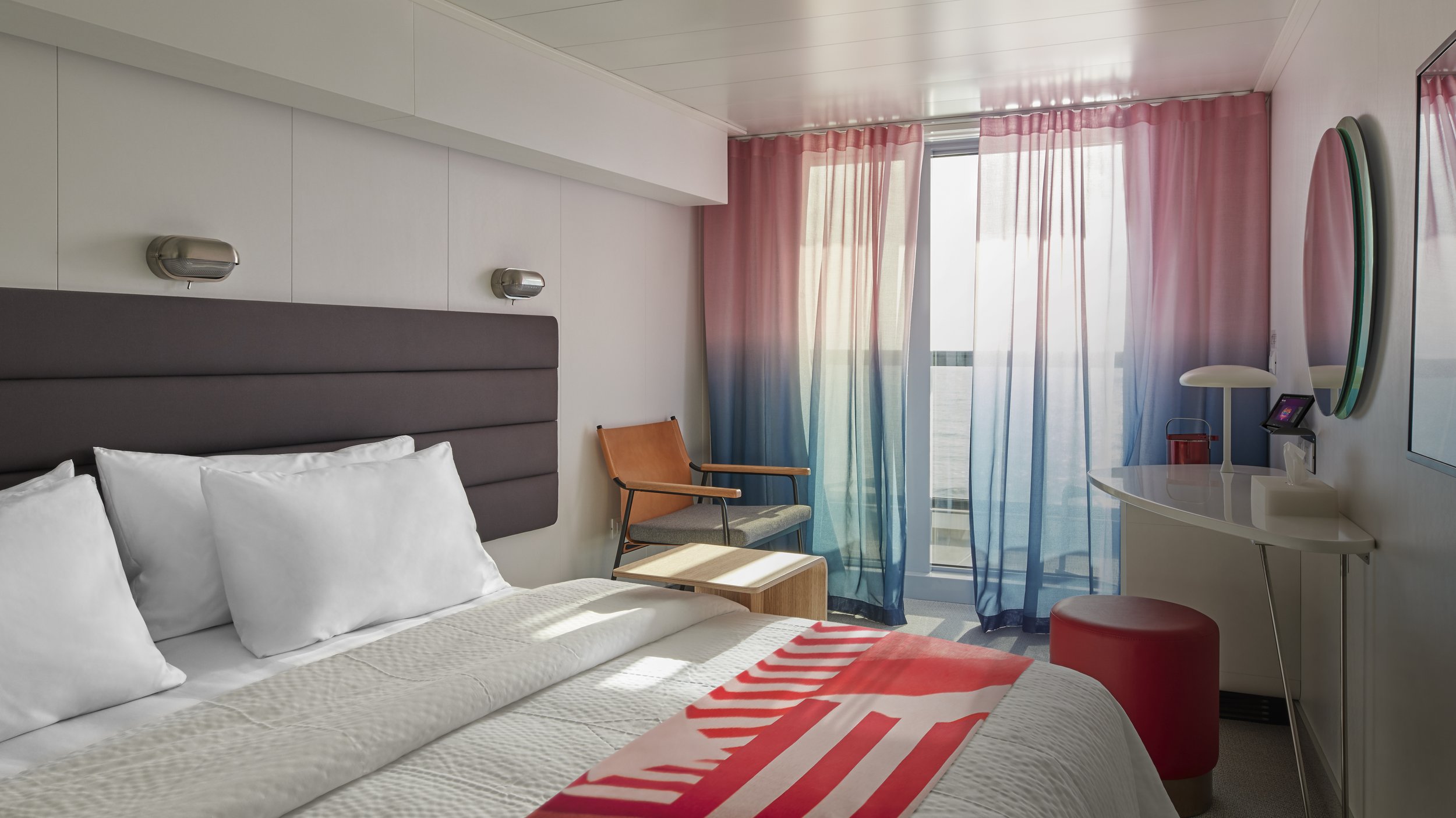 A hotel room with a bed, white pillows, a brown headboard, a wooden chair, pink and blue curtains, a white curved desk with a mirror, a small digital device, and a red round stool.