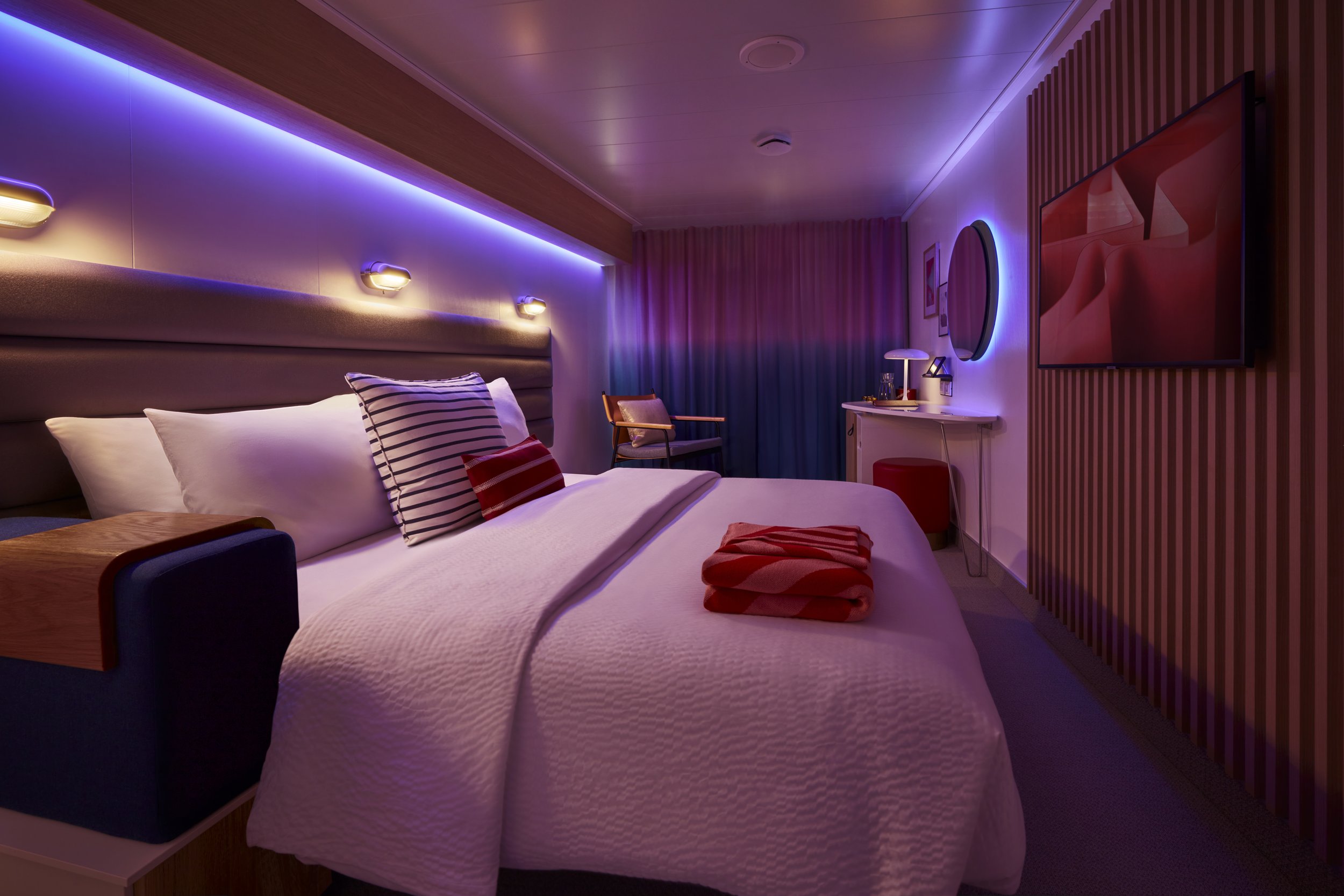 A modern hotel room with a bed, side table, wall-mounted TV, and cozy seating area, illuminated with purple and blue accent lighting.