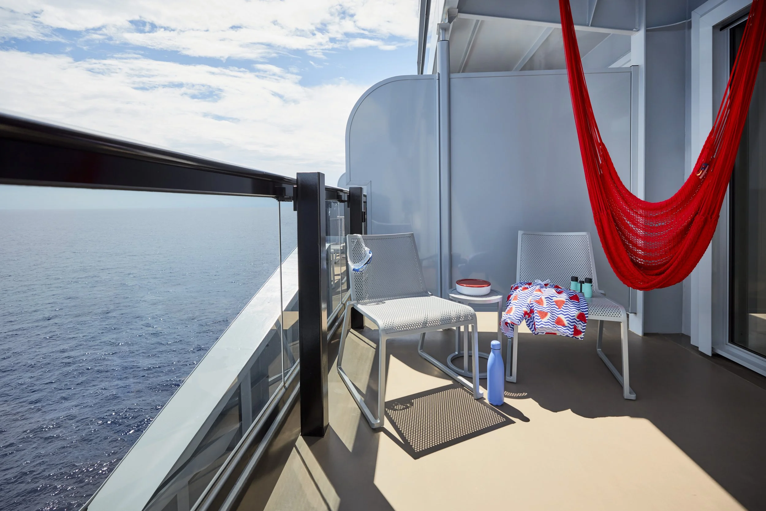 A balcony on a cruise ship overlooking the ocean with a blue sky and clouds, featuring two white chairs, a small round table with a bowl and a bottle, a red hammock, beach towel, and sunglasses.