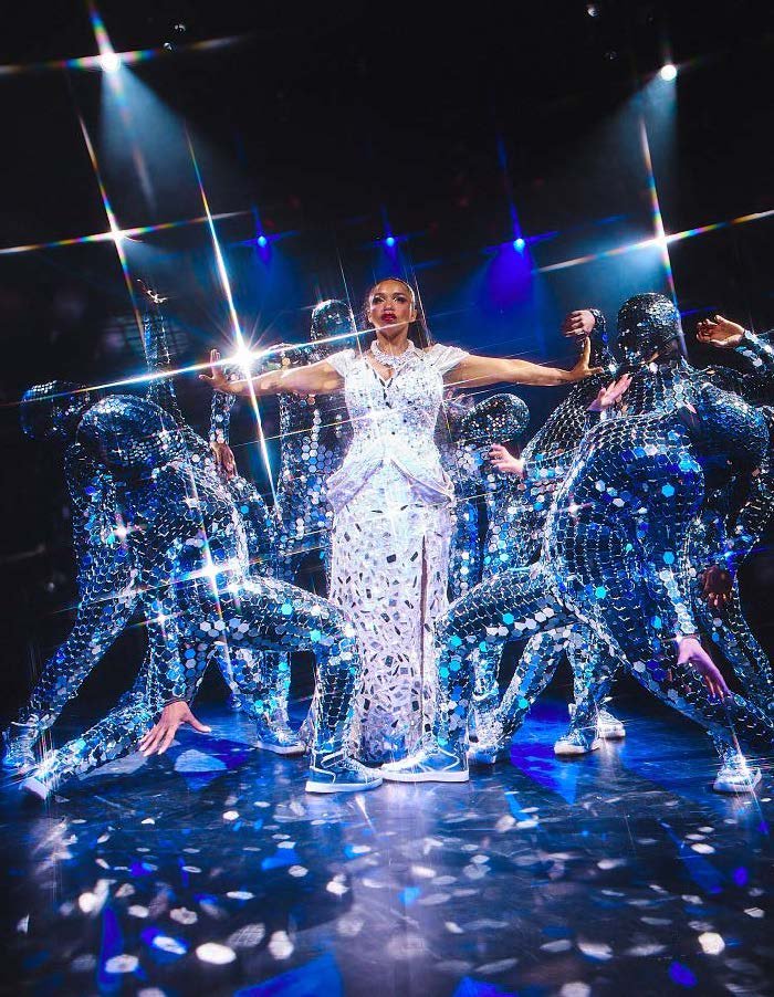 Performer in white outfit on stage surrounded by dancers dressed in mirrored costumes, with bright stage lighting and reflections creating a sparkling effect.