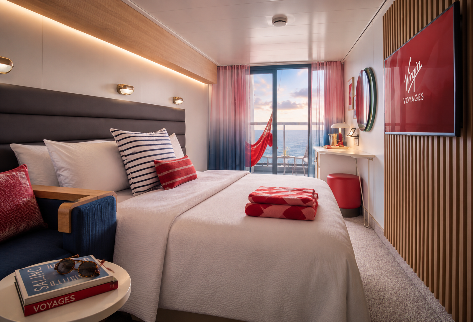 A modern hotel room with a large bed adorned with striped and solid red pillows, a view of the ocean through glass sliding doors, and a hammock outside on the balcony. The room features a flat-screen TV displaying 'Virgin Voyages', a desk with a lamp, and a small round table with reading glasses and books labeled 'Voyages' and 'Calling.'