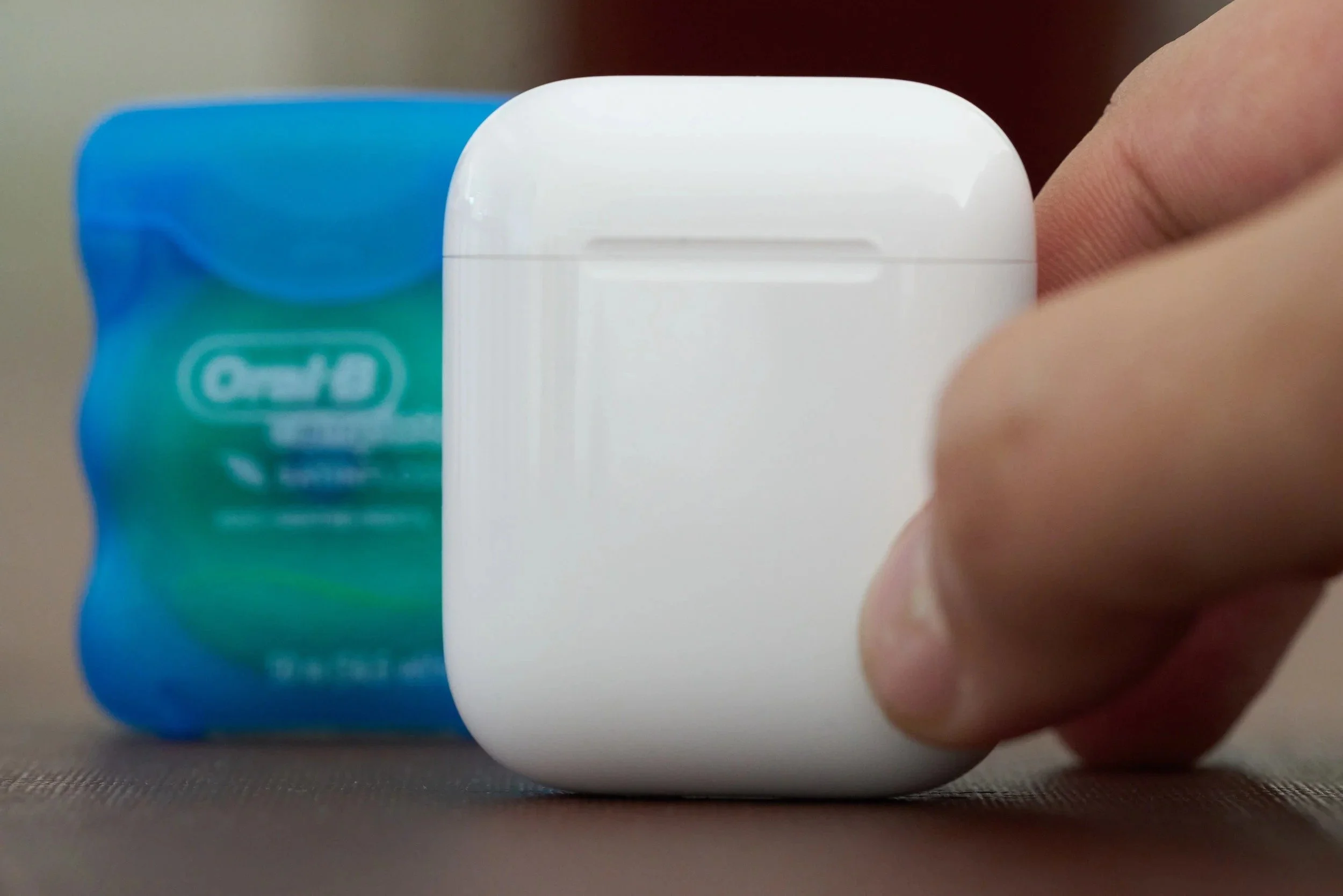 A hand holding a closed white AirPods charging case next to a container of dental floss, showing how small the case is.