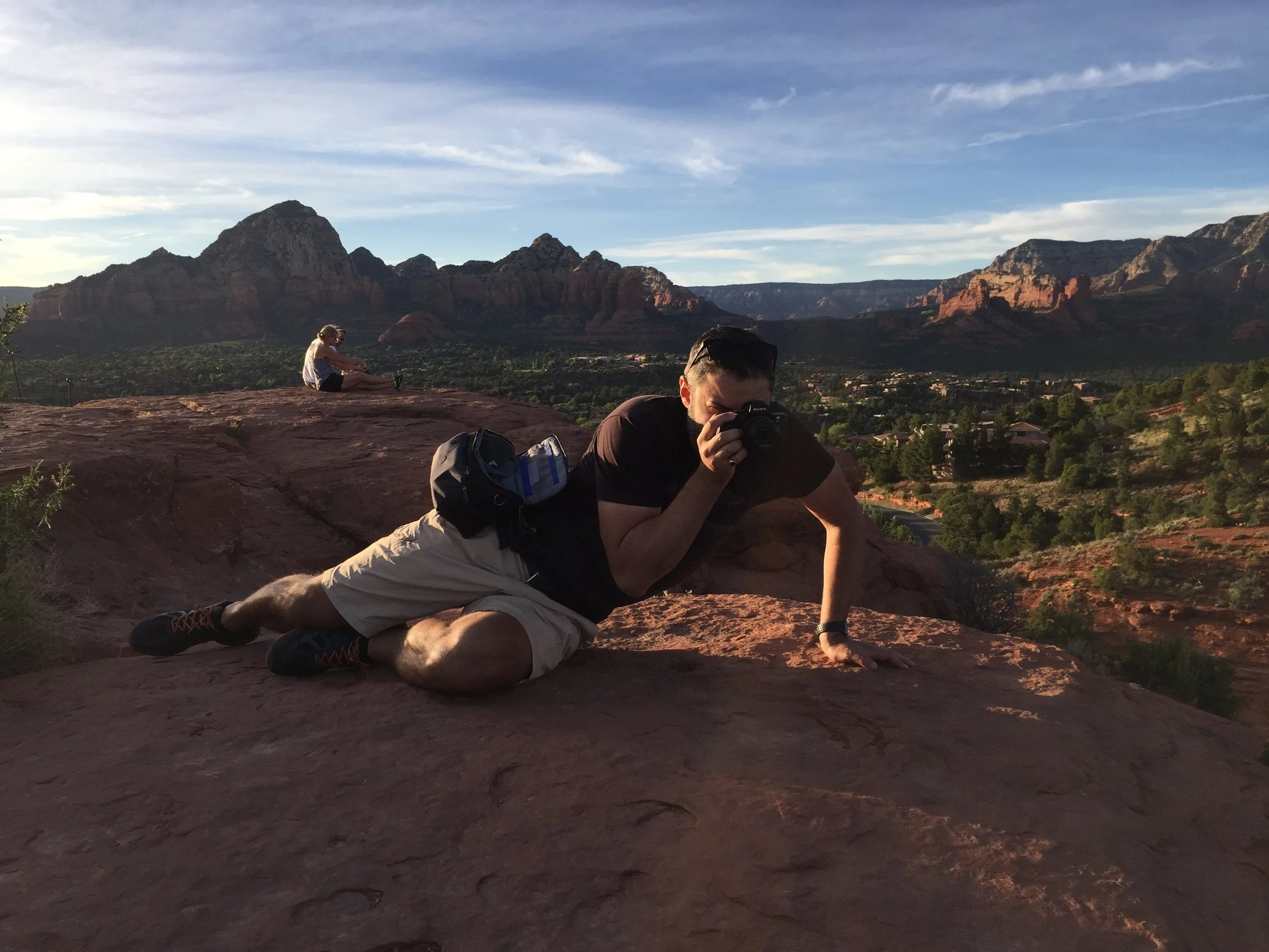 Eliyahu Ungar-Sargon lying prone on red rock above the Sedona valley, shooting with a camera in the late afternoon light.