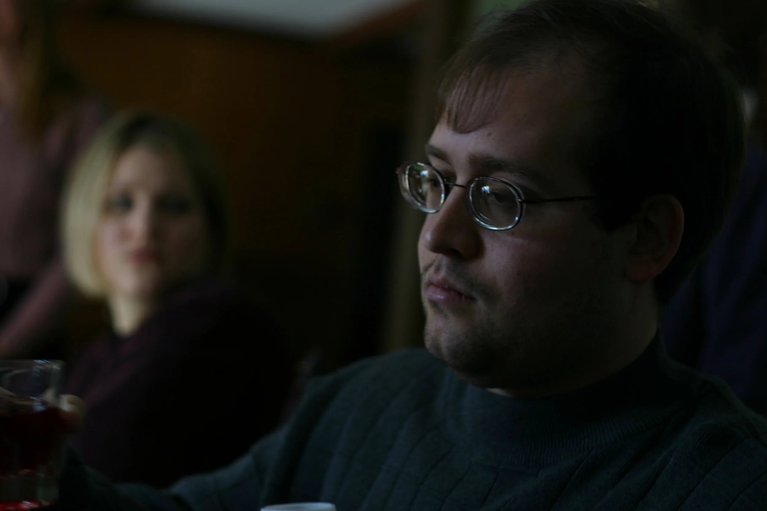 A man in glasses listens across a dinner table in low warm light, a woman blurred behind him.