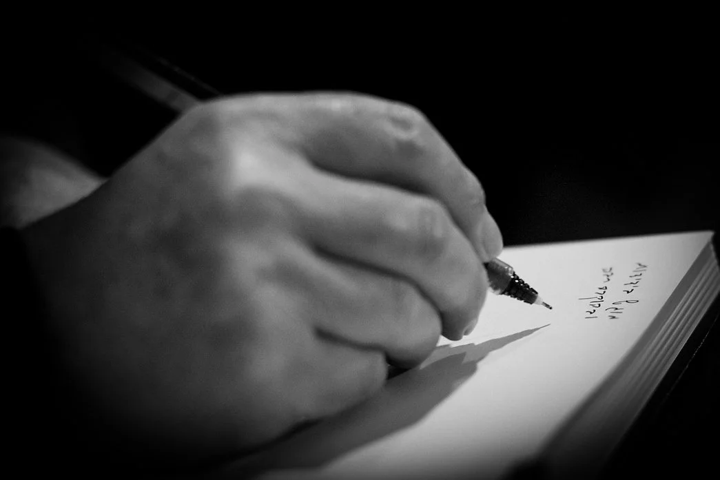 Black-and-white close-up of an elderly man's hand writing Hebrew script with a pen on a white page.