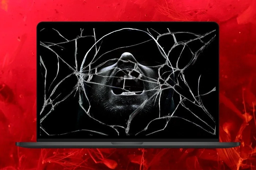 A laptop on a blood-red background, its screen showing a black-and-white photograph of a man shouting, seen through shattered glass.