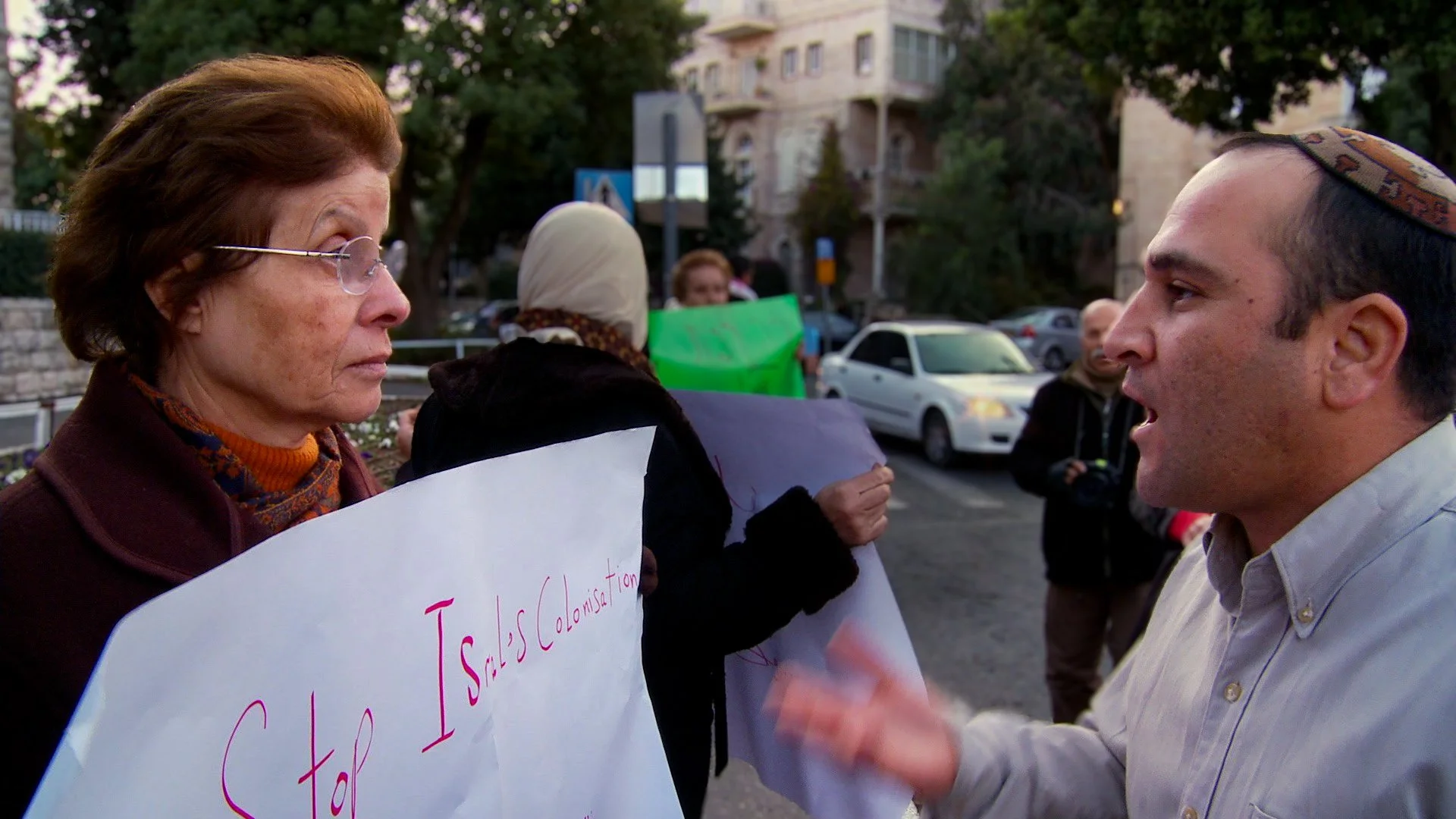 A Palestinian woman holding a protest sign and an Israeli man in a kippah argue face to face on a Jerusalem street.