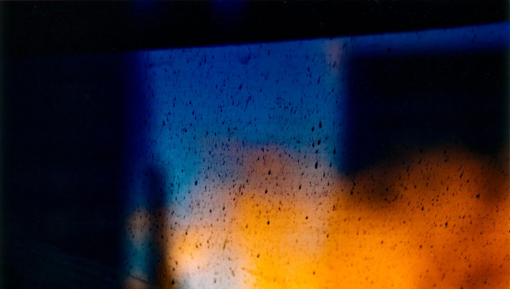 Abstract photograph of deep blue and amber light glowing through rain-speckled glass in darkness.