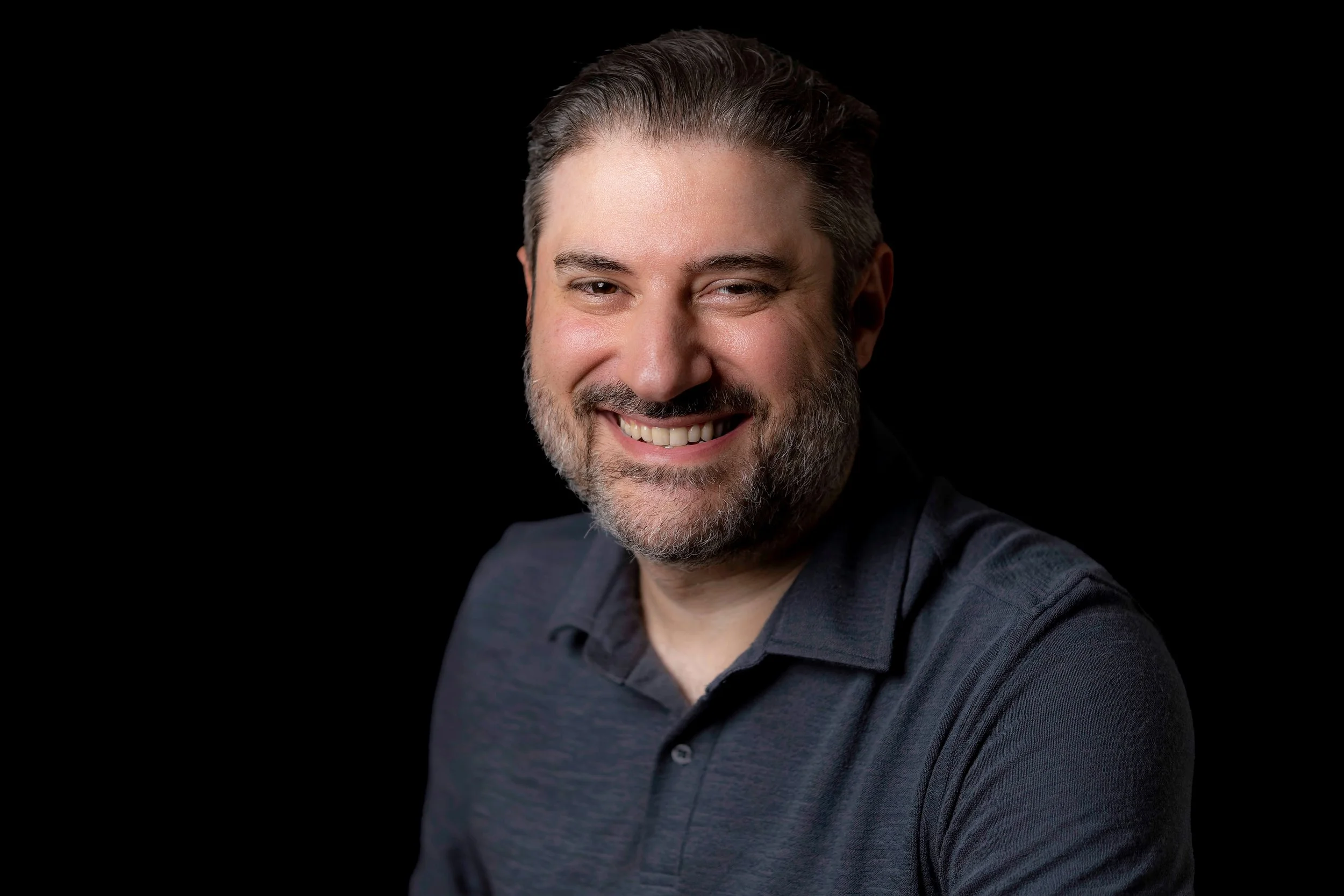 Portrait of Eliyahu Ungar-Sargon smiling, lit from the side against a black background.