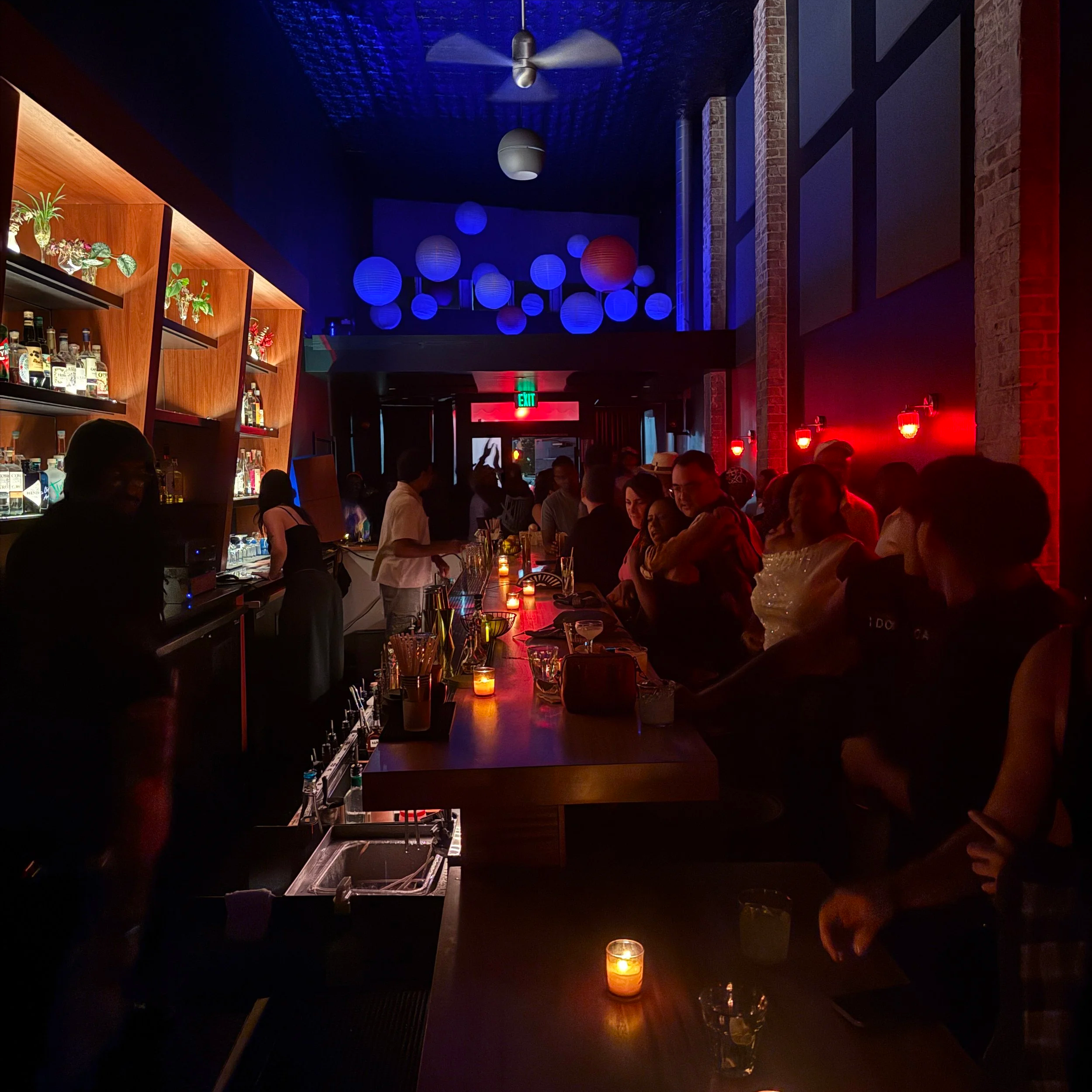 Wide shot of Moonglow from the rear, showing off the tall ceilings, exposed brick columns and soft red lighting.. The room is full of smiling guests