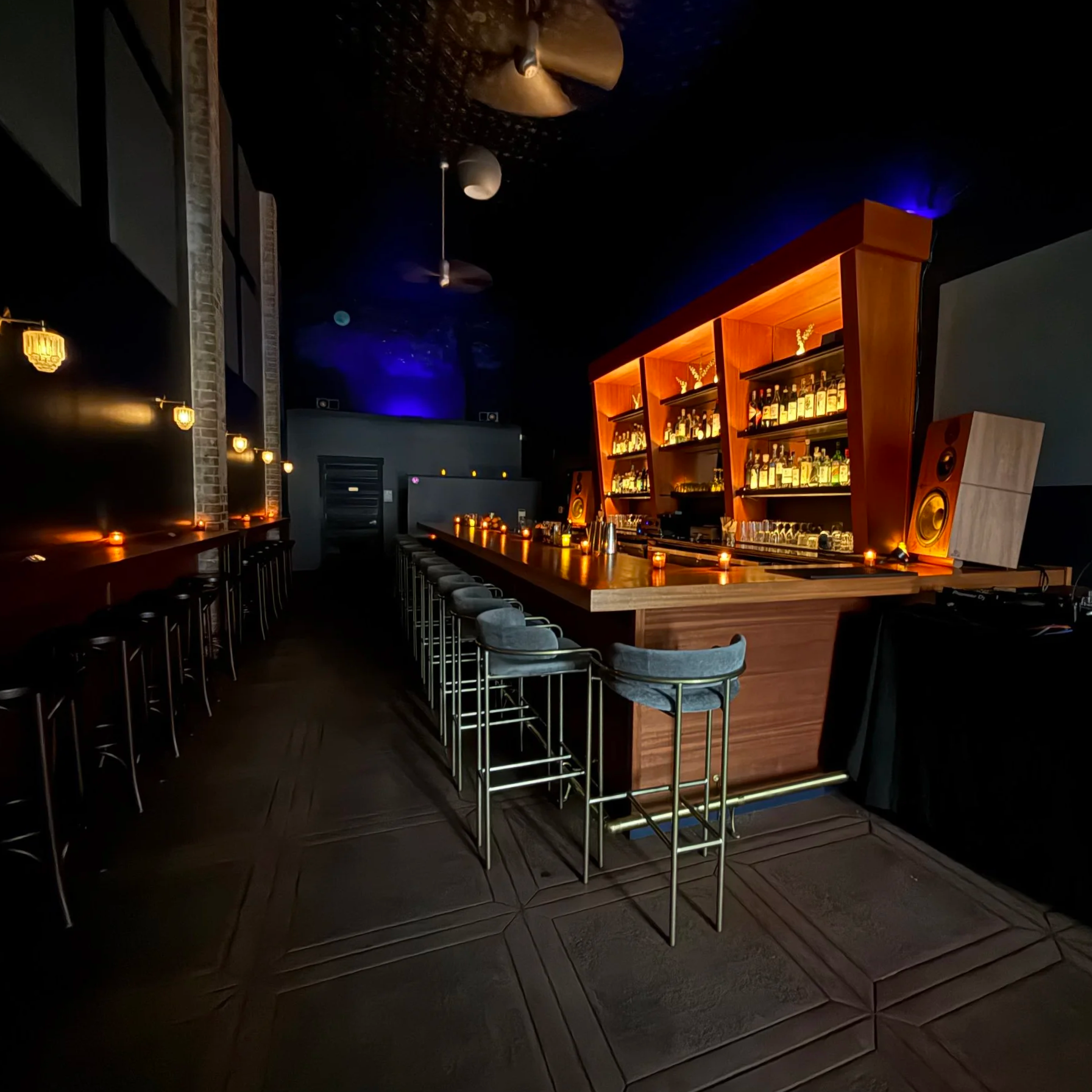 Wide shot of a dimly lit, Art Deco-inspired bar. An angular, wooden back bar shelf has illuminated liquor bottles on it and the long mahaogony bar is accented with glowing light.
