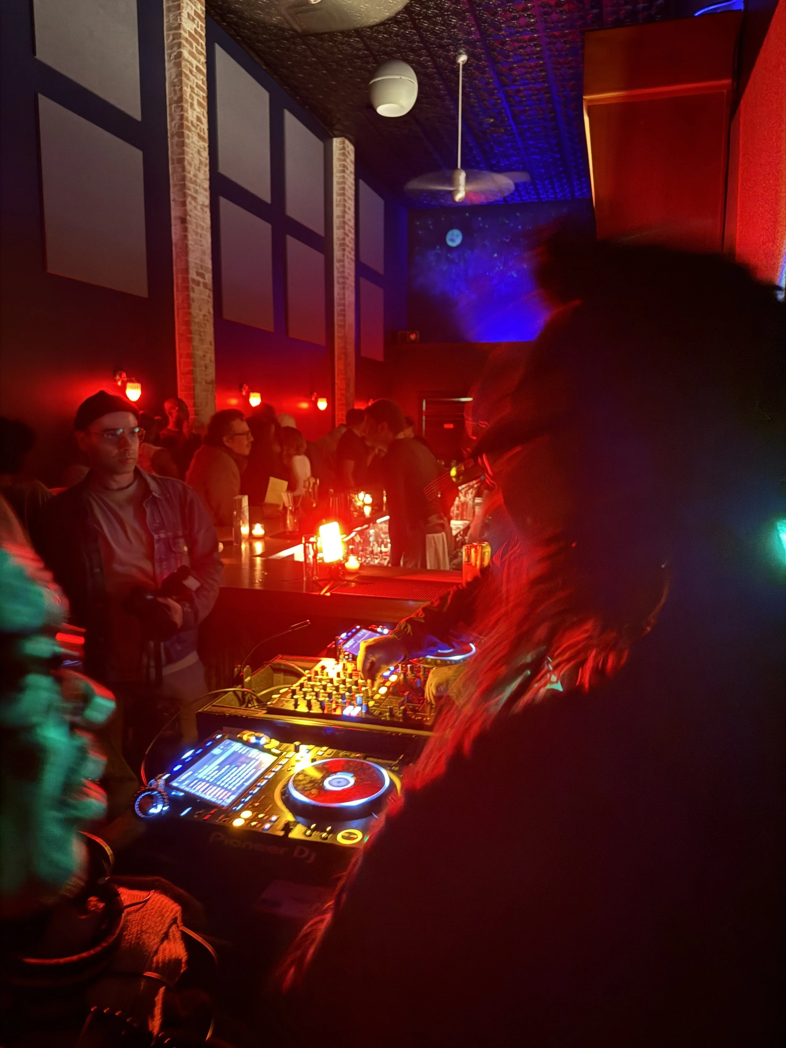 Glowing red lights illuminate a crowded bar, in the foreground a DJ plays on CDJs and adjusts the EQ knobs on a mixer.