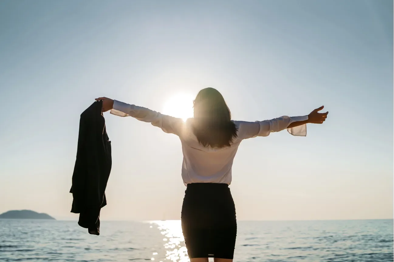 Woman standing by the ocean with arms outstretched and holding a jacket, backlit by the sun, at sunrise or sunset.