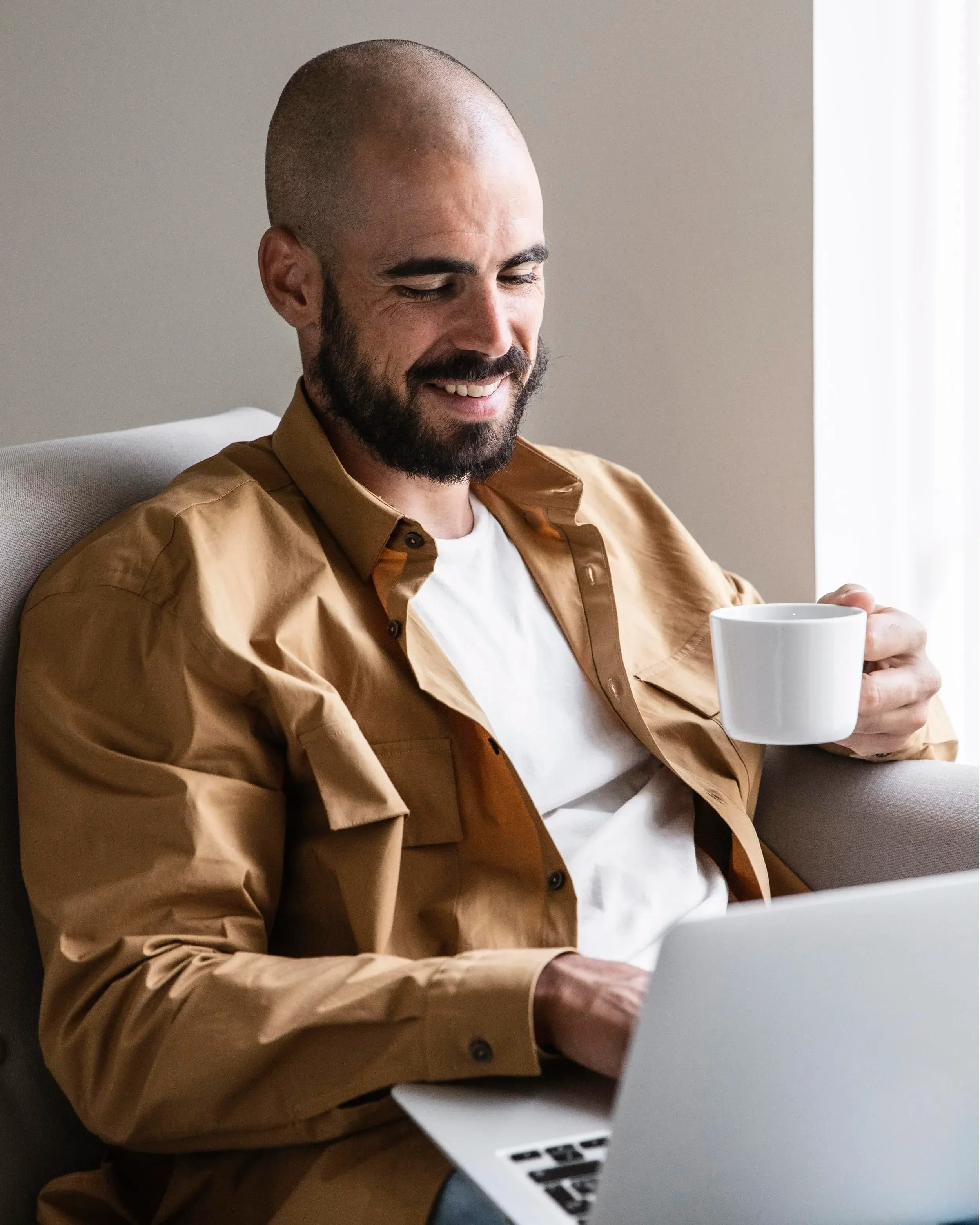 A man with a bald head and beard sitting at a desk, smiling while holding a white mug and looking at a silver laptop.