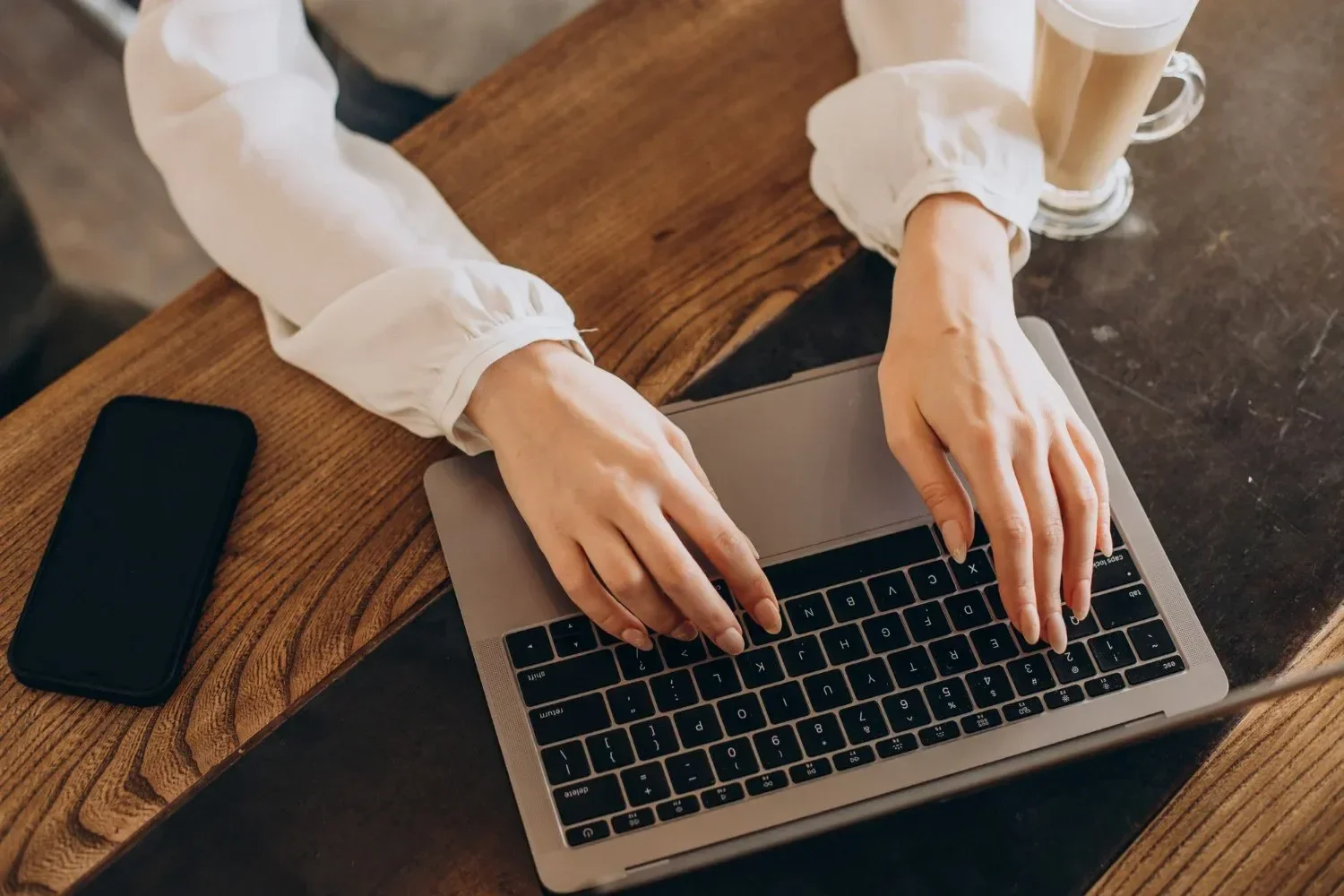 Person typing on a laptop at a wooden table, with a smartphone nearby and a latte in a glass mug.