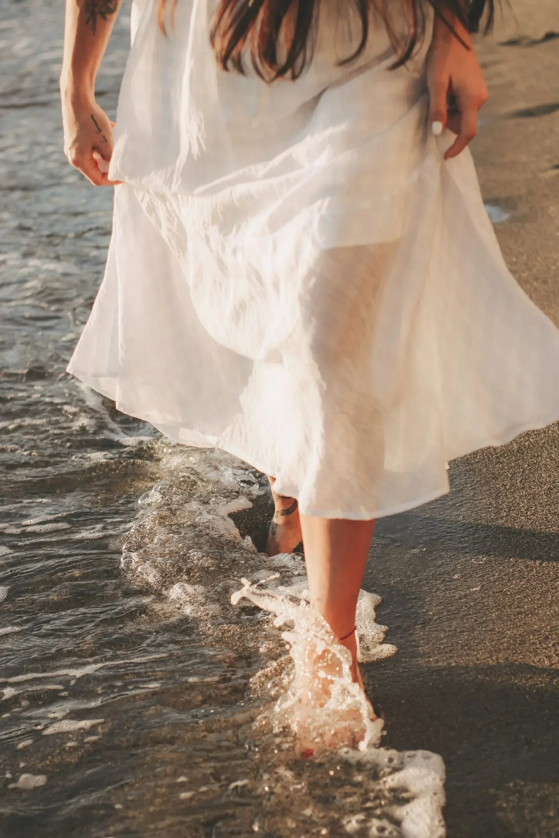 A person wearing a white dress walking in the ocean, with the waves touching their feet and the sunlight reflecting off the water.