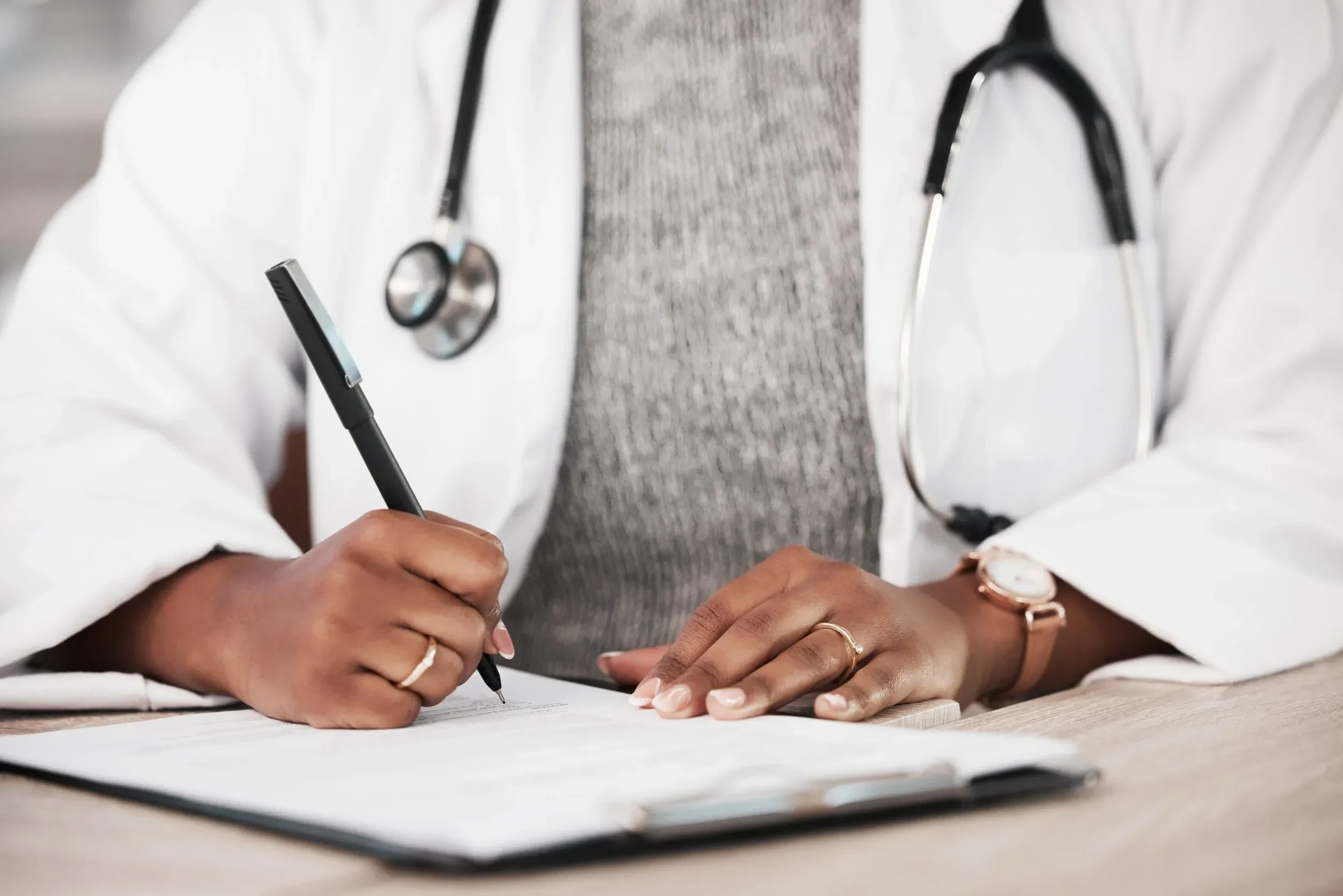 A healthcare professional wearing a white coat with a stethoscope around their neck writing on paper with a pen, and a person wearing a watch and rings resting their hands on the paper.