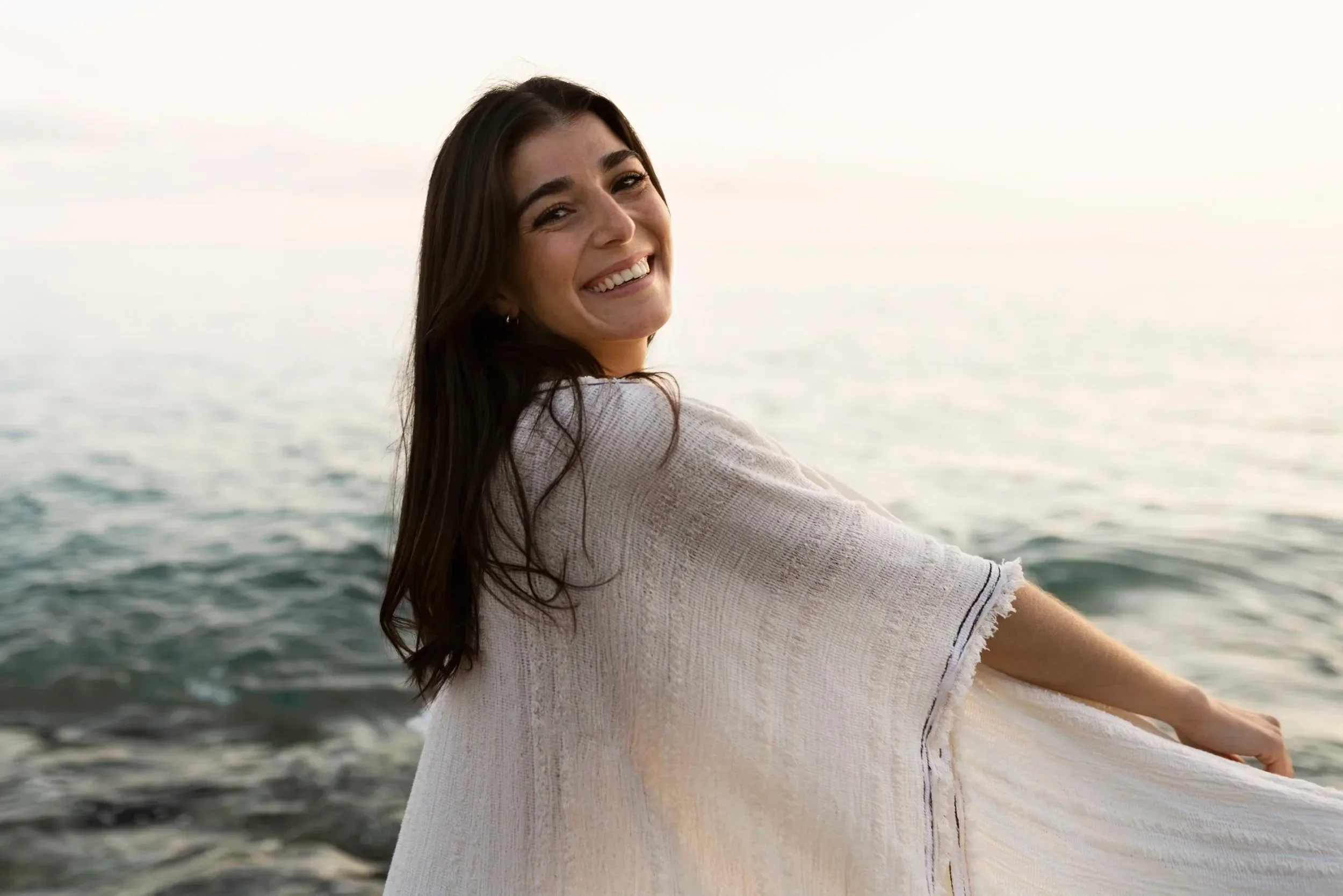 A woman with long dark hair smiling at the beach, wearing a beige sweater, with ocean waves in the background.