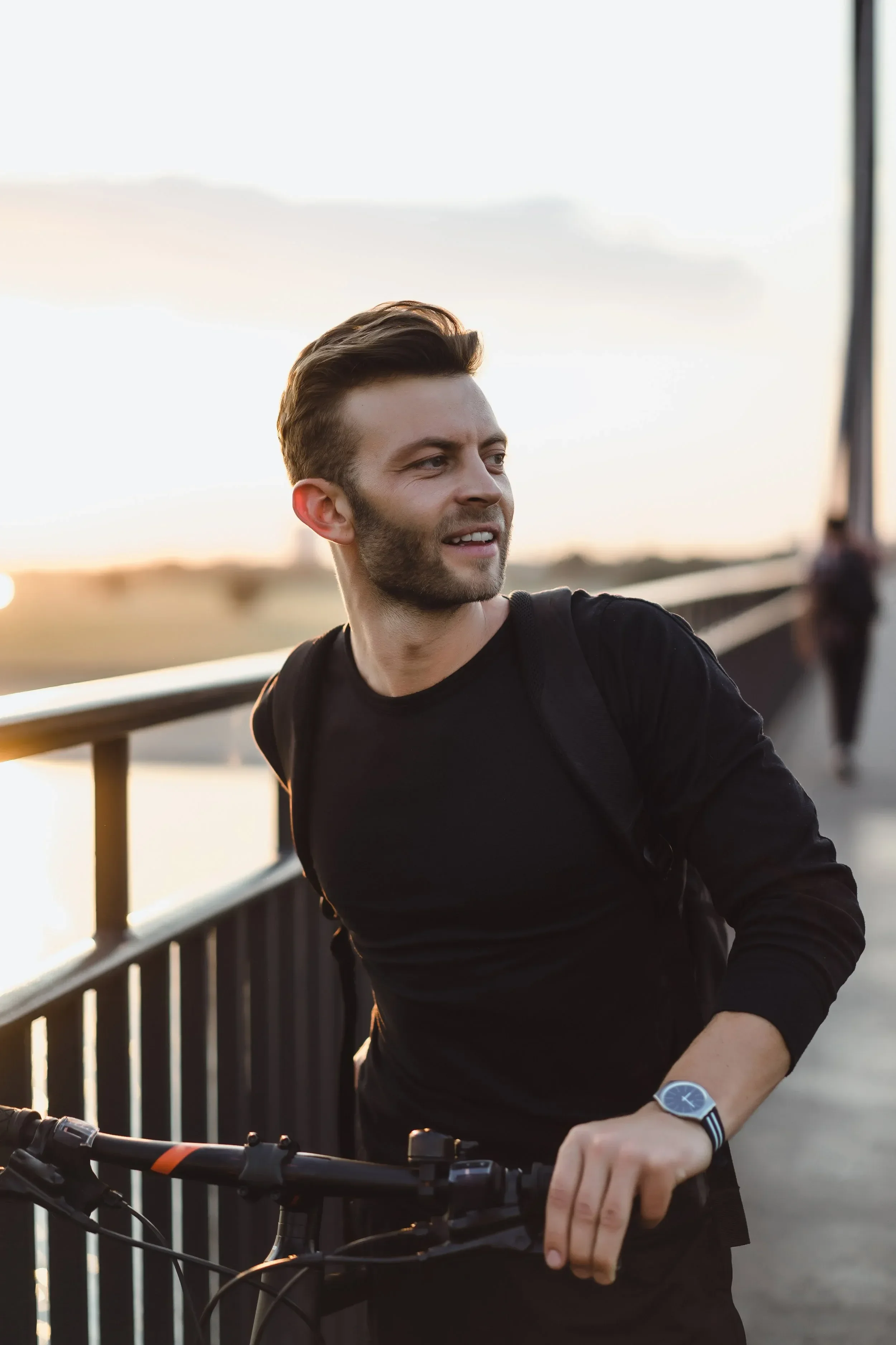 A man with styled brown hair and a beard, wearing a black long-sleeve shirt and a watch, is standing outdoors near a railing during sunset, holding a bicycle. The background is slightly blurred with a calm sky and distant landscape.