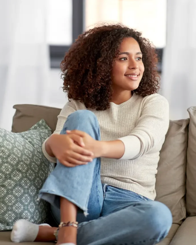 A young woman with curly hair sitting on a couch, smiling and looking to the side, wearing a beige sweater and blue jeans.