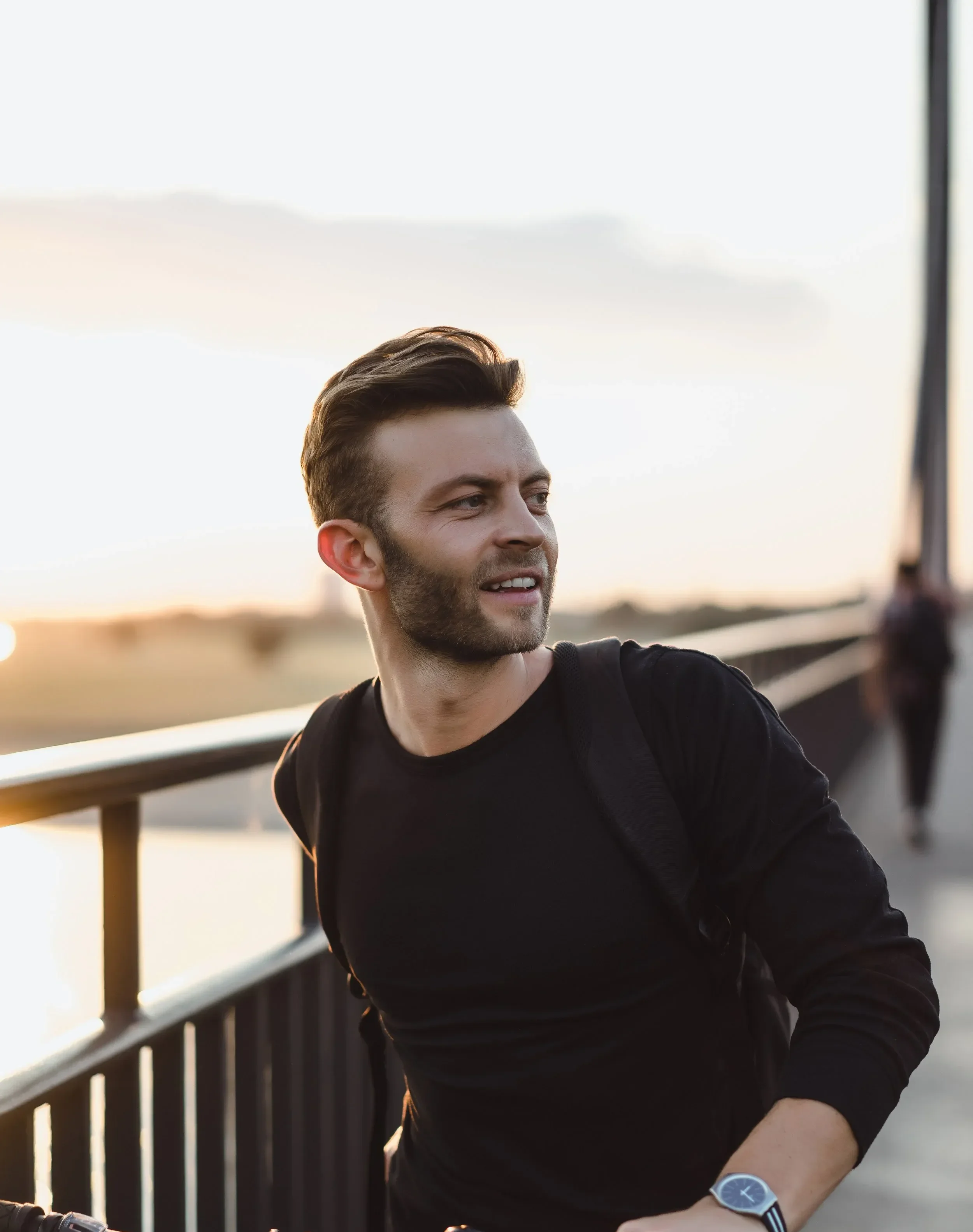 A man with styled brown hair and a beard, wearing a black shirt and a blue wristwatch, standing outdoors on a bridge during sunset.