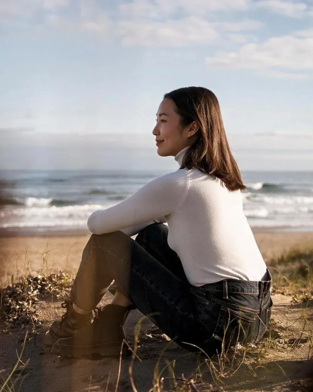 A woman sitting on the beach, facing the ocean, with a relaxed and content expression, wearing a white top, black pants, and black shoes, surrounded by sand and dunes under a partly cloudy sky.