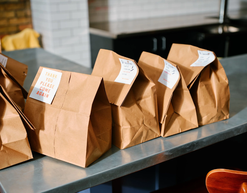 Several brown paper takeout bags on a countertop in a kitchen, with a sticker that says 'Thank you, please come again'.