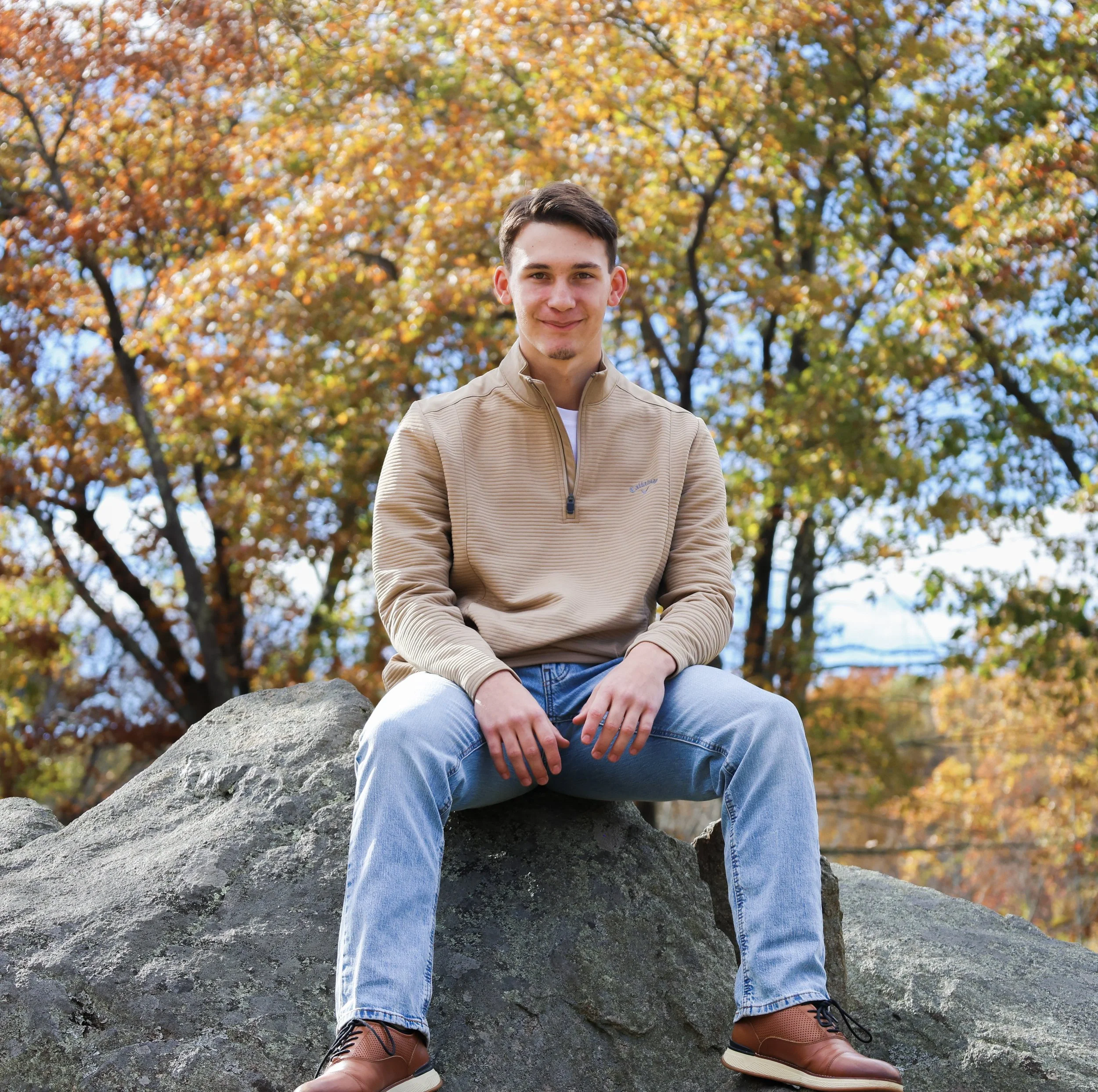 A young man is sitting on a large rock outdoors during fall, surrounded by trees with colorful autumn leaves. He is smiling, wearing a beige pullover, blue jeans, and brown boots.