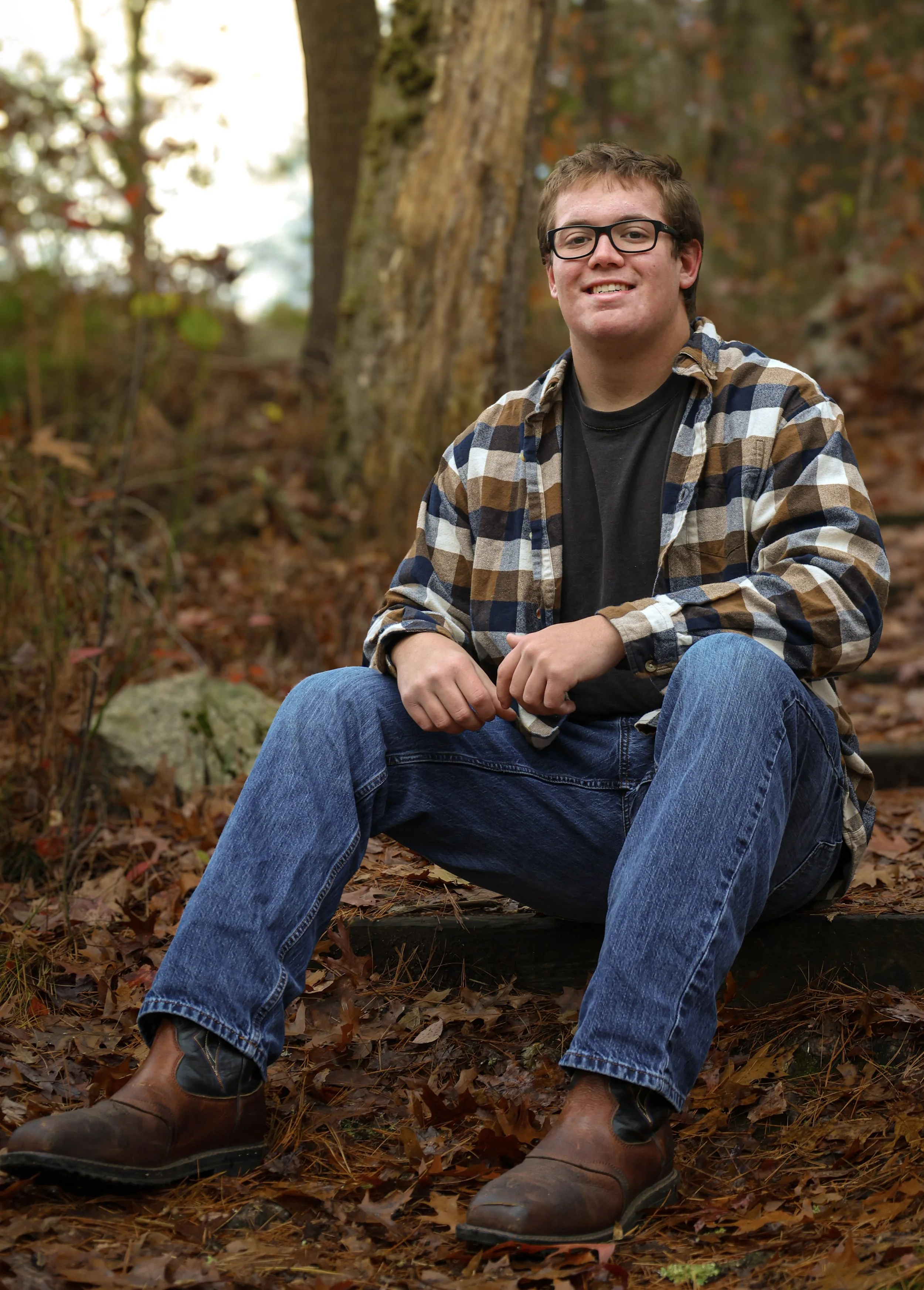 A young man with glasses, wearing a black shirt, plaid shirt, blue jeans, and brown boots, sitting outdoors on the ground amidst fallen autumn leaves with trees in the background.