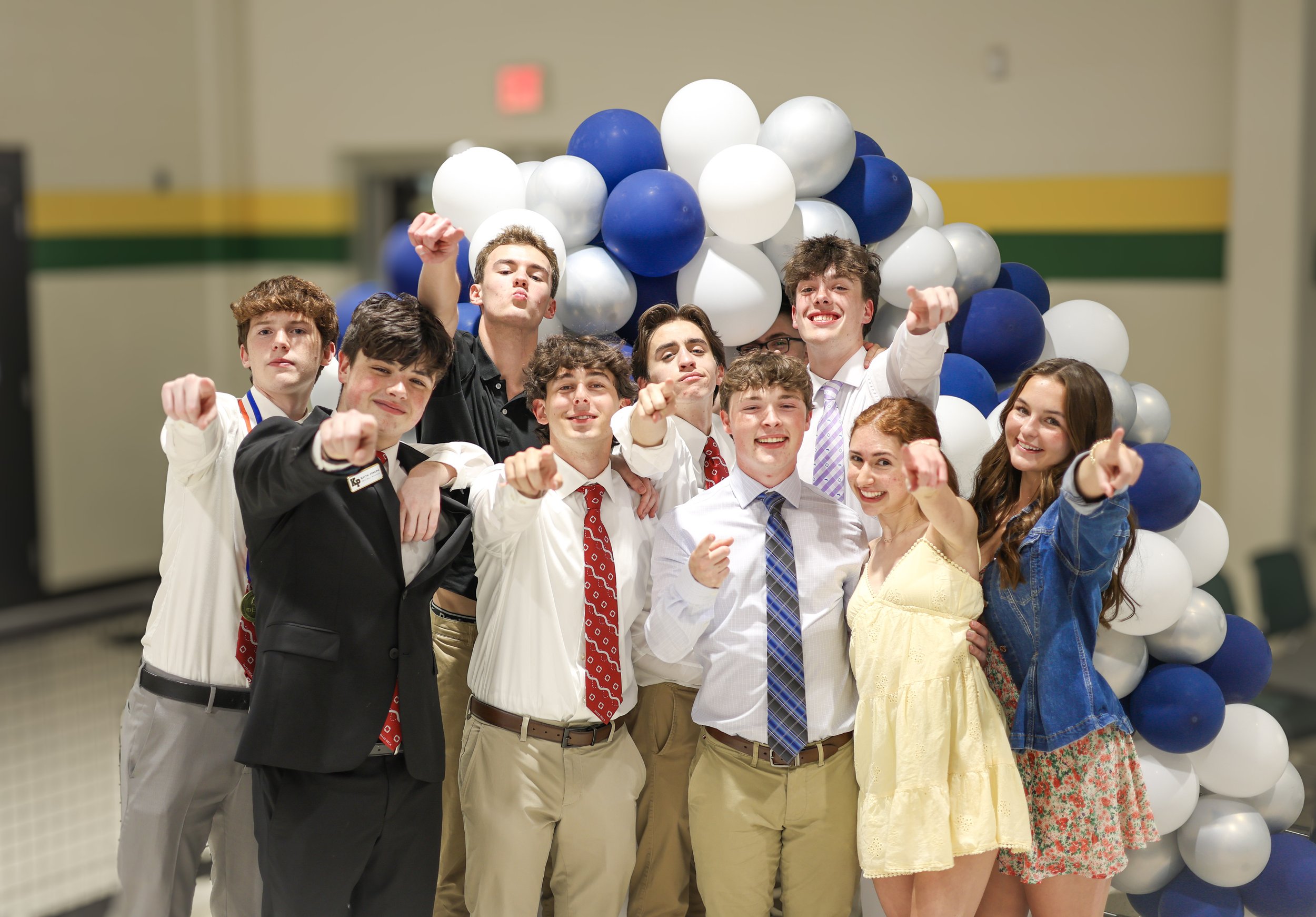 A group of young people celebrating indoors with balloons, some pointing at the camera and smiling.