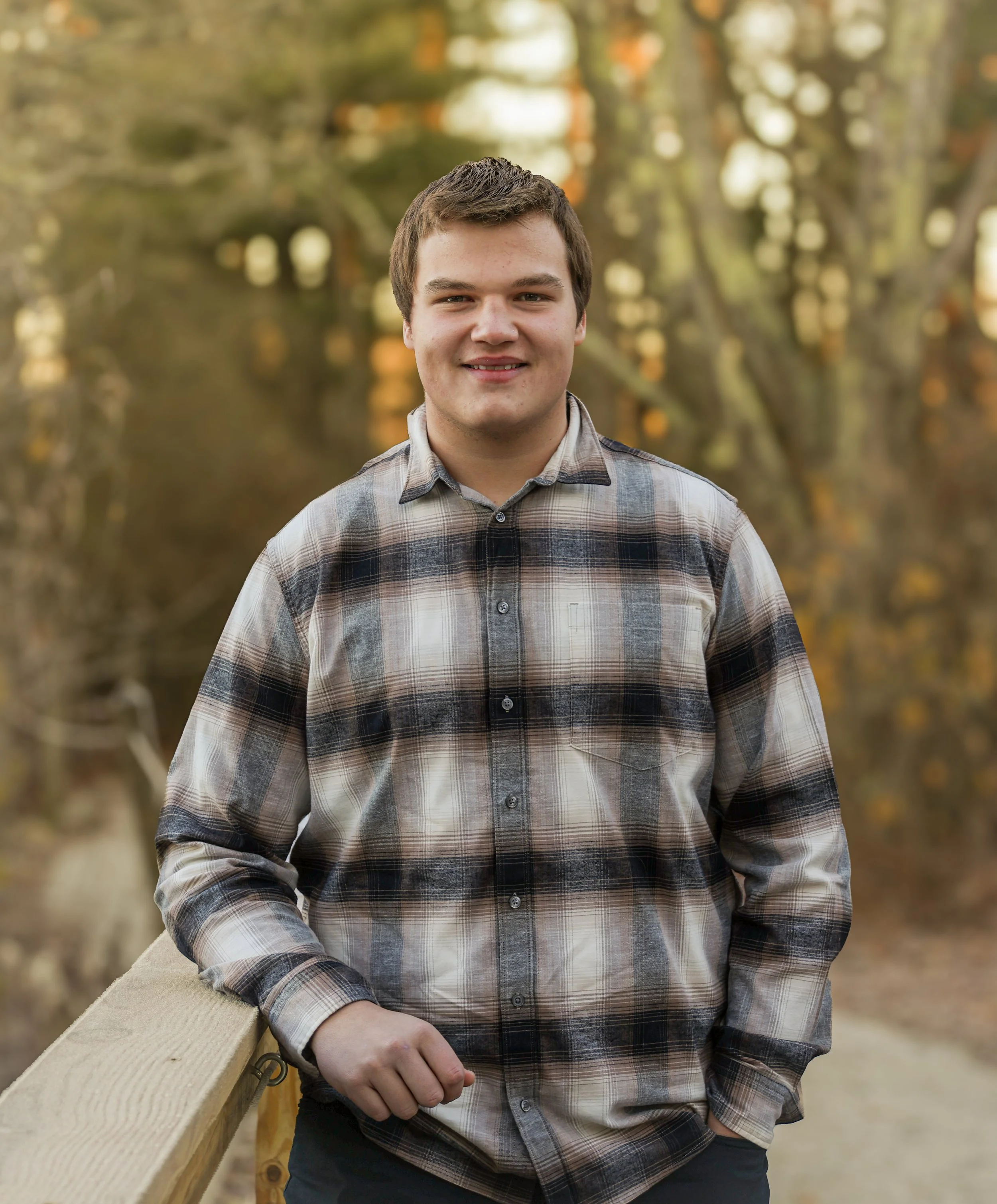A young man with short brown hair and a light beard, wearing a plaid shirt, standing outdoors during fall. He is leaning against a wooden railing in a park with blurred trees and autumn leaves in the background.