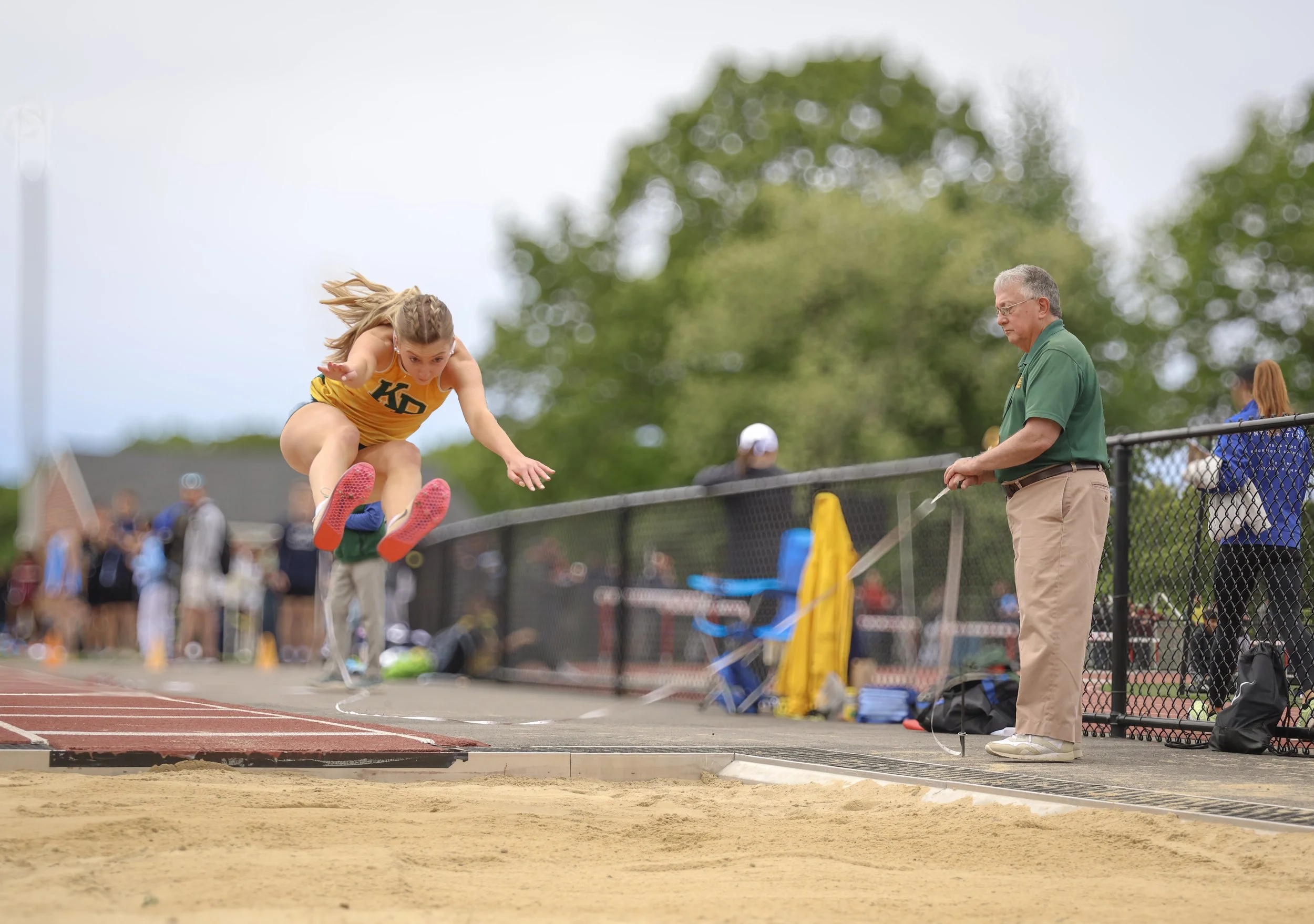 A young female athlete in a yellow track uniform jumping into a sandpit for a long jump event during a track and field competition, with an official in a green shirt holding a measuring tape nearby, and spectators blurred in the background.
