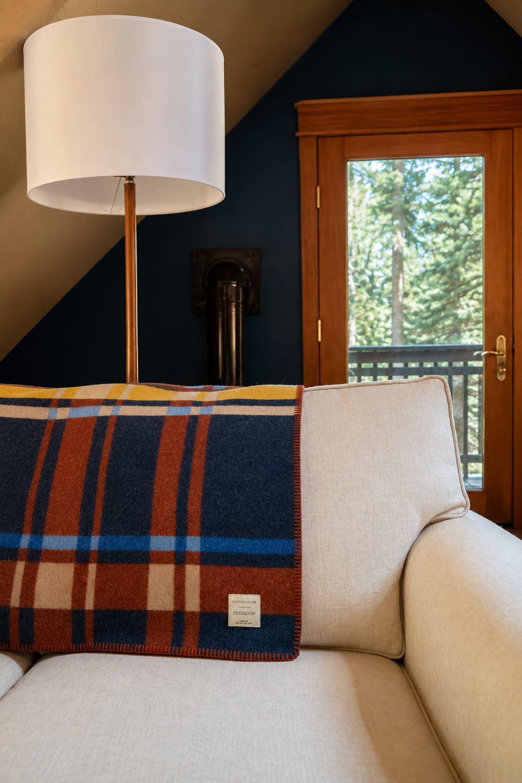 A cozy living room with a beige sofa, a plaid blanket draped over the back, a white modern floor lamp, and a wooden-framed door leading outside to trees.