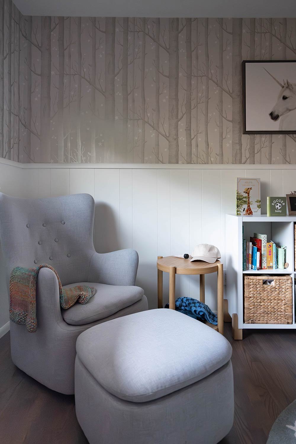 A cozy corner with a light gray upholstered armchair and matching ottoman, a small wooden side table with a baseball cap, bookshelves filled with children's books, a framed picture of a white unicorn, and wall with forest-themed wallpaper featuring trees and stars.