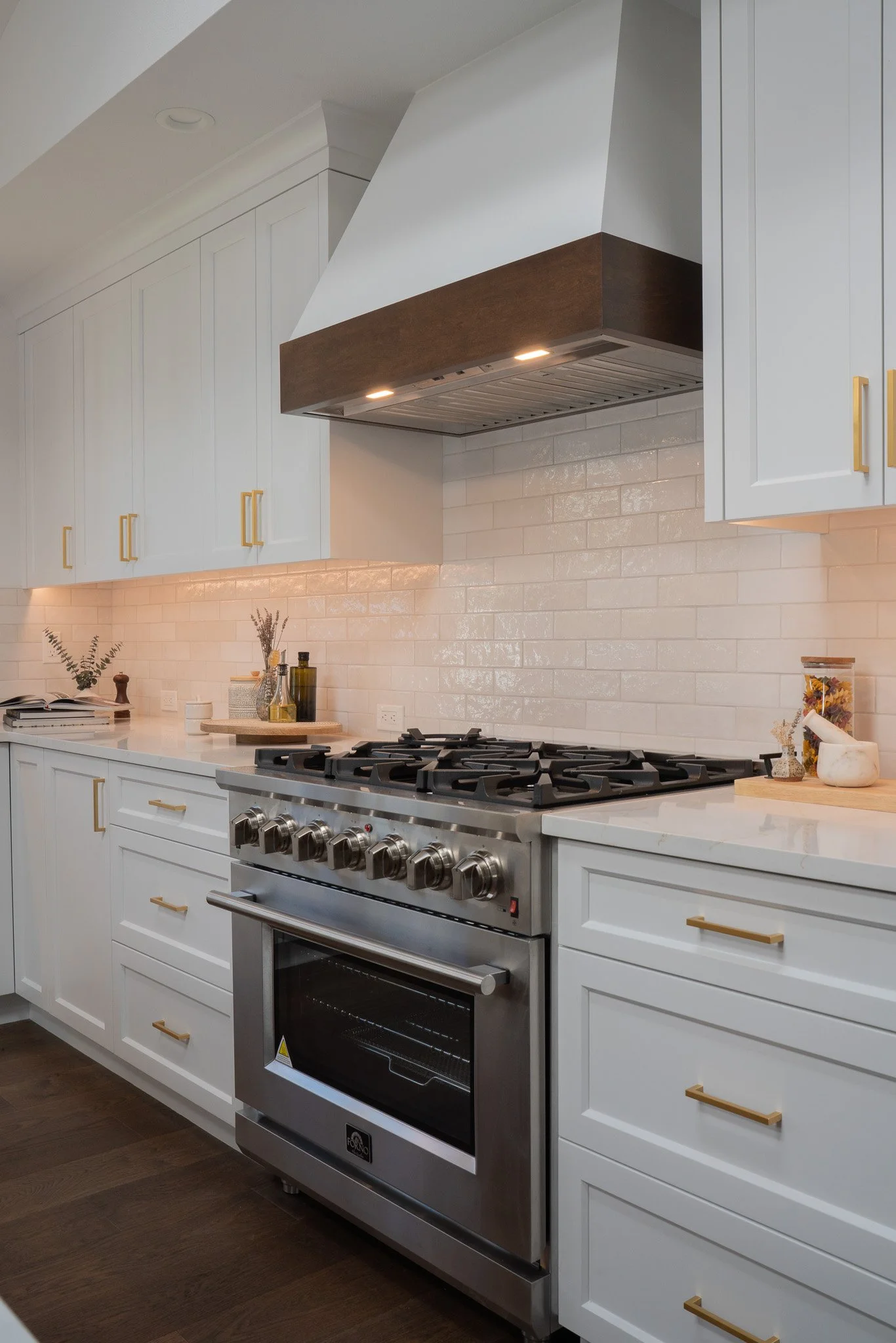 A modern kitchen with white cabinets and gold handles, a stainless steel gas oven, and a white subway tile backsplash. On the countertop, there are decorative items including a small potted plant, salt and pepper shakers, and jars of spices.