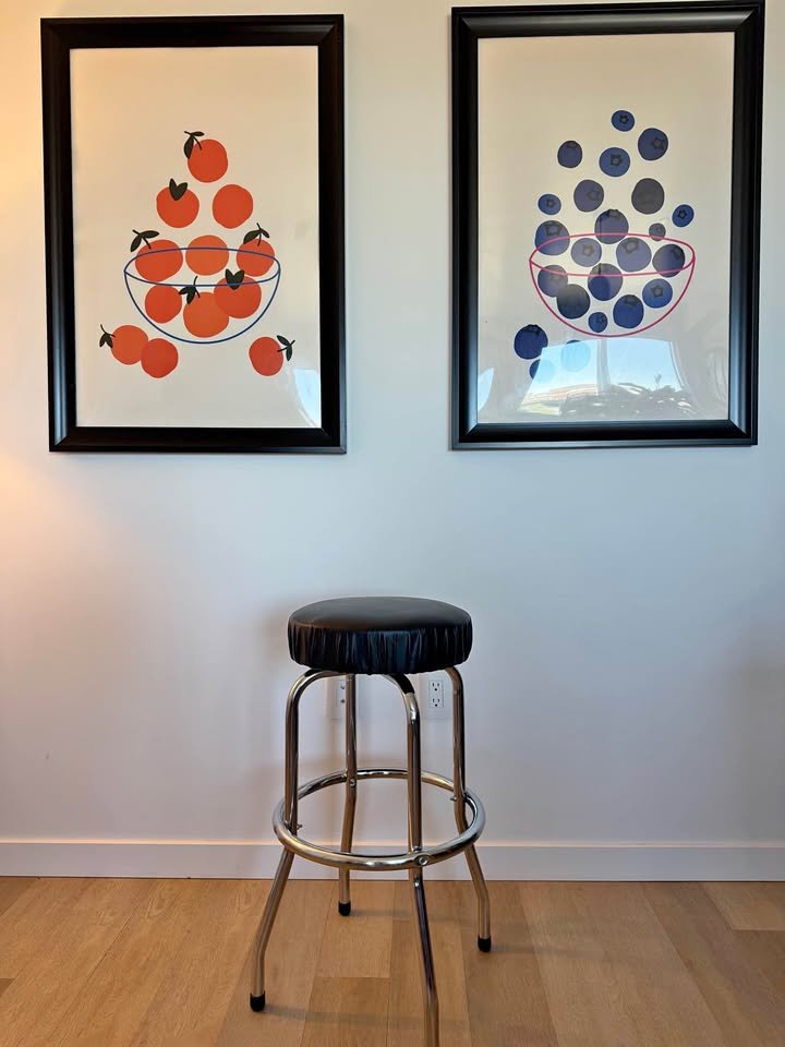 Two framed art prints of fruit, one with oranges and the other with blueberries, hanging on a white wall above a black and chrome barstool.