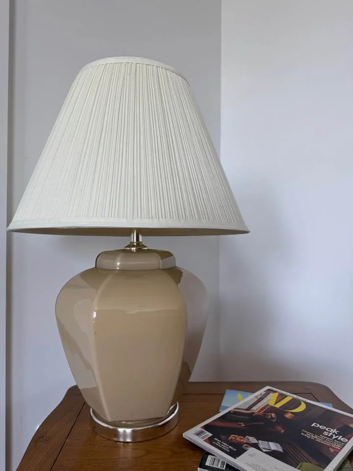 A beige ceramic table lamp with a white pleated lampshade placed on a wooden surface next to magazines.