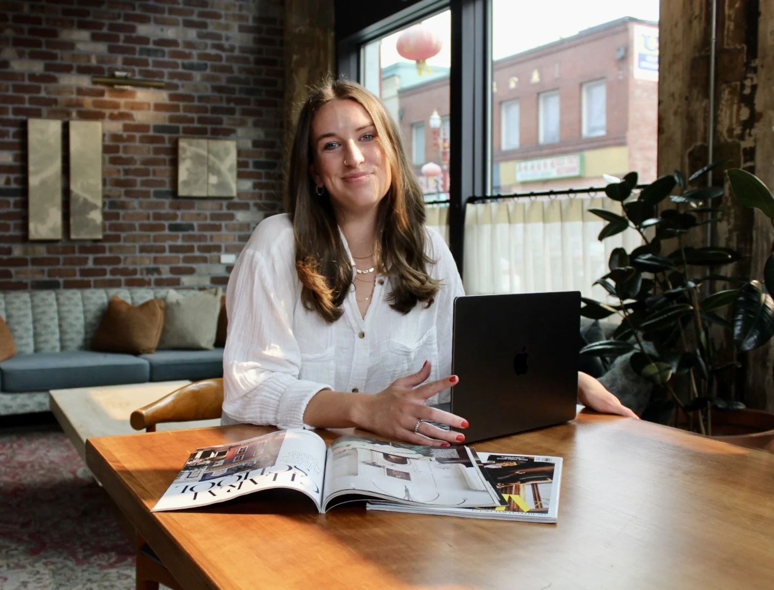 A woman with long brown hair and wearing a white blouse sitting at a wooden table with a black laptop and magazines, in a cozy cafe with brick walls, large window, plants, and sofa in the background.