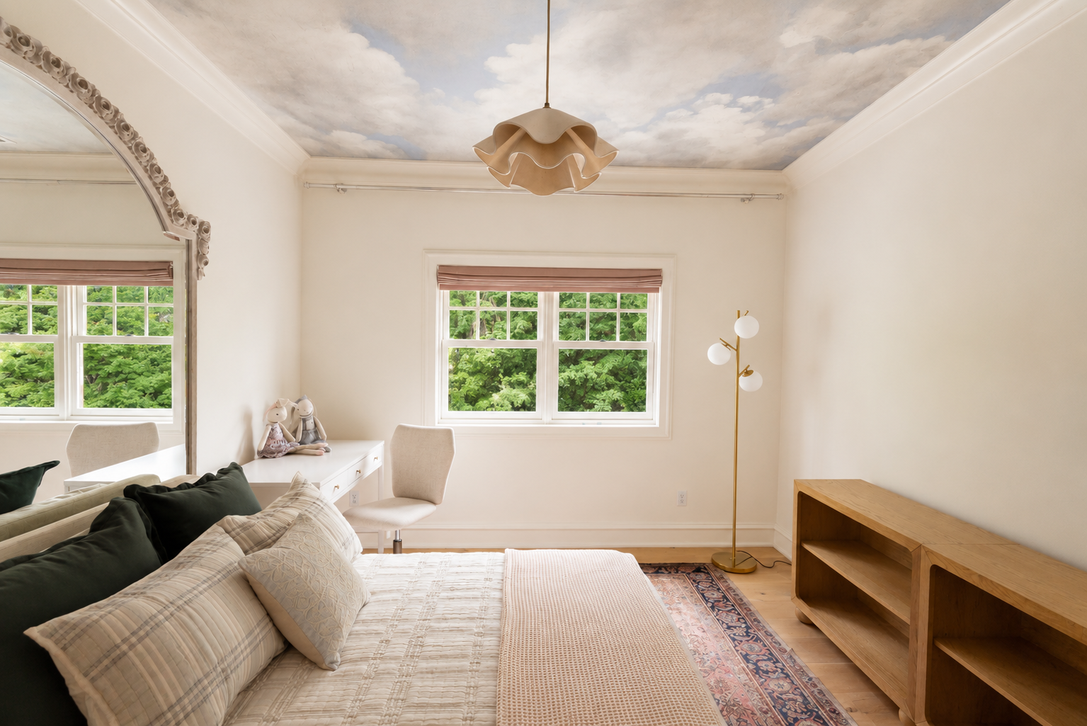 A bedroom with light-colored walls and a ceiling painted to look like a cloudy sky. There is a window with greenery outside and a white desk with plush toys inside. The bed has cream and dark green pillows. A wooden bookshelf is on the right, and a m