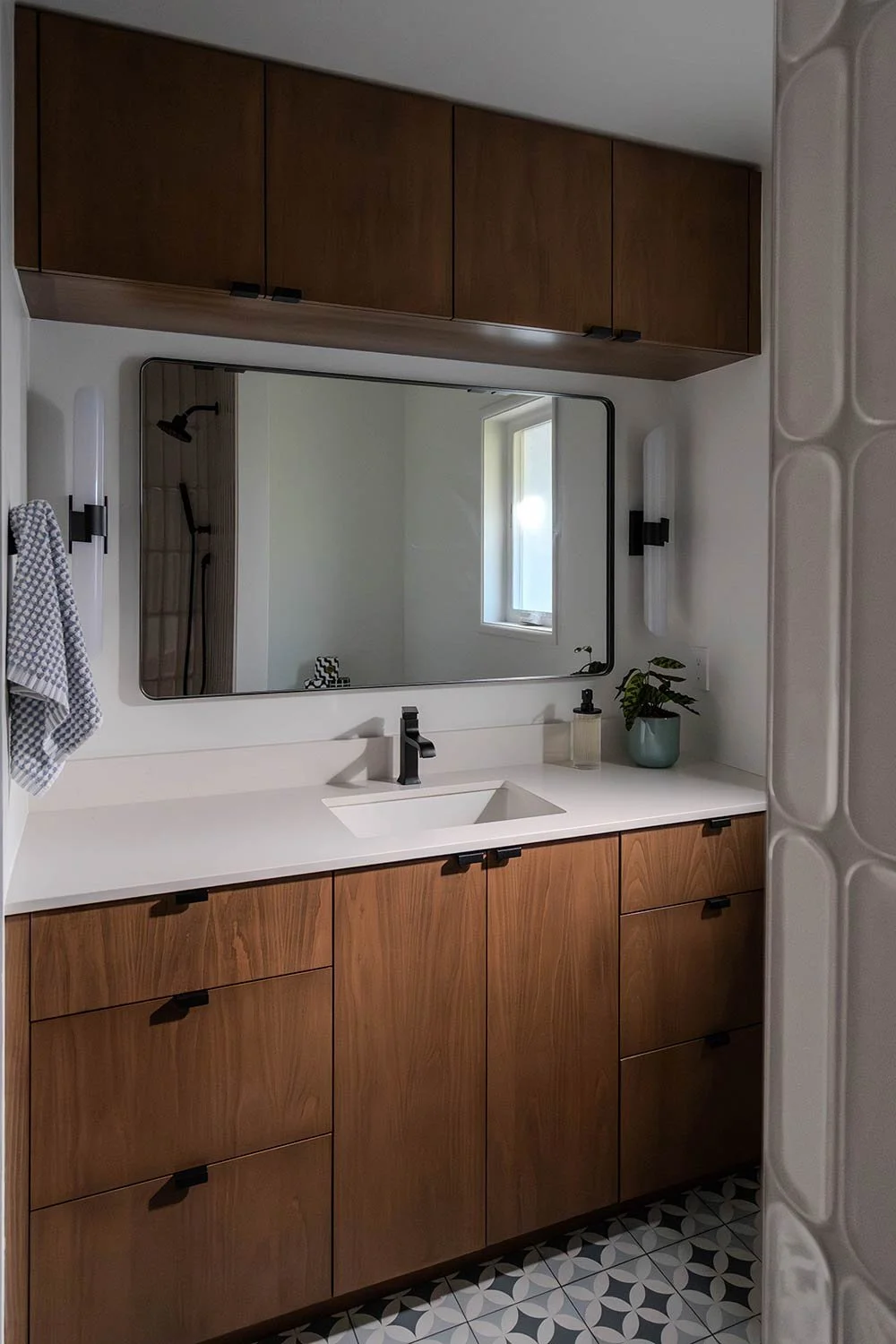 Modern bathroom with wood cabinets, a large mirror, black fixtures, plants, and geometric patterned floor tiles.