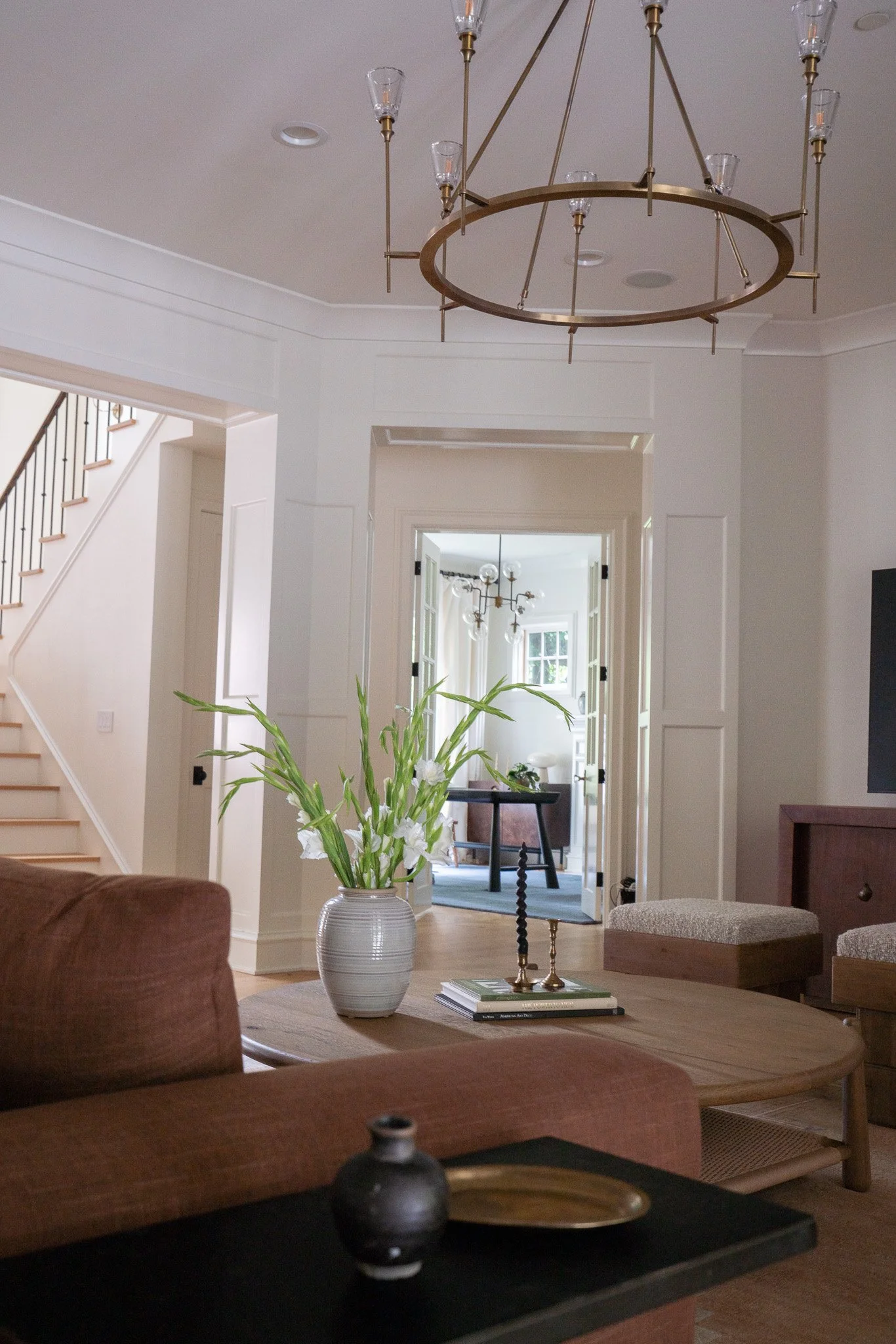 A view of the interior of a home highlighting a chandelier hanging from the ceiling, a hallway with open doors leading to a room with a pool table, and a table with books and a brass candle holder, with a green plant in a white vase in the foreground